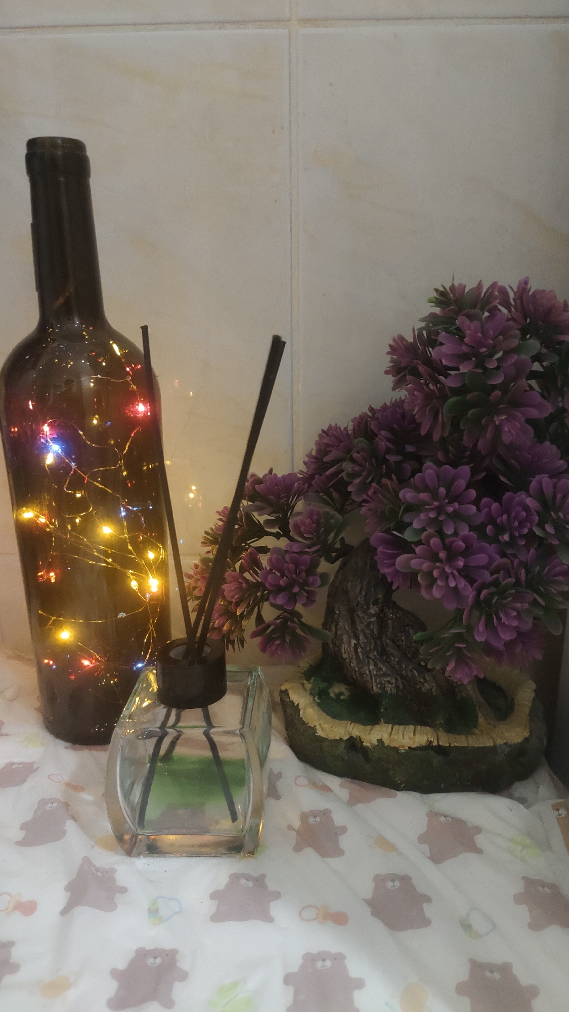 An indoor still life scene, likely from Krasnoyarsk, Russia, features a collection of decorative and functional household items arranged against a backdrop of light-colored, possibly tiled, walls.
In the left foreground, a tall, dark glass bottle stands upright, illuminated from within by a string of multi-colored fairy lights, casting red, yellow, and blue glows. To its right, a clear glass diffuser bottle contains several dark reeds, suggesting an air freshener or essential oil setup. Further to the right, a vibrant artificial plant resembling a bonsai tree displays numerous clusters of small, purple flowers or foliage, with a textured brown trunk and a base covered in green and brown material.
All these objects rest on a light-colored fabric surface, which appears to be patterned. The pattern consists of repeating motifs of cartoonish brown bears, interspersed with small, colorful pacifiers and possibly other baby-related illustrations, suggesting it could be a baby blanket, changing mat, or similar textile. The overall arrangement creates a cozy and decorative ambiance. No specific activity is taking place, and there is no visible text. The indoor setting, combined with the illuminated bottle, implies the image might have been taken in the evening or a dimly lit space.