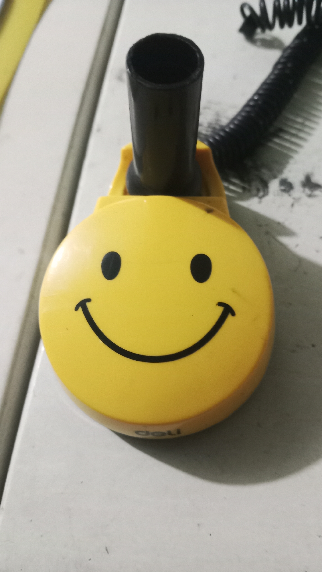A yellow, circular object with a smiley face design is positioned on a white, textured surface. The object has two black oval eyes and a black curved line for a mouth. A black tube extends vertically from the top center of the yellow object. To the right of the yellow object, a black coiled cord is visible, lying on the same white surface. The background is out of focus, but suggests a vertical surface to the left and a textured surface behind the objects. The lighting creates a shadow to the right of the smiley face object. This scene is not located in Keraton, Indonesia.