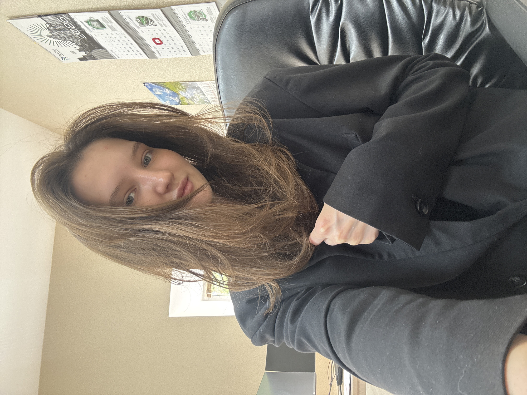 A young woman with long, light brown hair and light-colored eyes is captured in a close-up, casual selfie, likely taken indoors in Perm, Russia. She is seated, looking directly towards the camera with a neutral expression, and her hair partially covers her face on the right side. She wears a black formal jacket or blazer over what appears to be a black hoodie or casual top.
The setting is an indoor space, possibly an office or study, with light beige or cream-colored walls. Behind her, a black faux-leather office chair is visible. On the wall above and to her left, several calendars are displayed. One prominent calendar features the year "2020" in large text and includes images that appear to be industrial or agricultural, along with the text "Neo-Tex". Other calendars show month names and dates, with visible Cyrillic script, such as what looks like "НОЯБРЬ" (November) and "ДЕКАБРЬ" (December). Below the calendars, a smaller picture of the Earth from space is visible. To her right, a window allows natural daylight into the room, and some faint green foliage can be seen outside, suggesting trees or bushes. What seems to be a desk or table and an electronic device are present in the lower right portion of the frame. The time of day appears to be daytime due to the natural light.