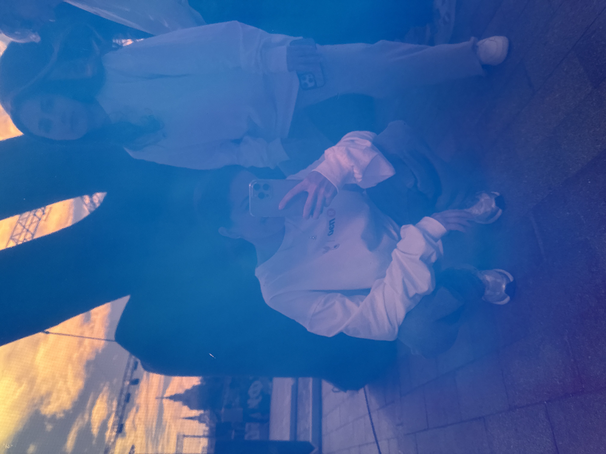 The image shows a reflection of two people, a woman and a child, in a large, dark, reflective surface, likely a window or polished material. The woman is in the foreground, taking a selfie with her phone. She is wearing a white hoodie and dark pants. The child is in the background, also wearing a white hoodie and light-colored pants, looking towards the camera.
The reflection includes a blurry background showing what appears to be a sunset or sunrise over a body of water, with architectural elements that could be from Moscow. The sky is filled with orange and yellow hues, casting a warm glow that is somewhat diffused by the reflective surface. The overall lighting suggests it is either early morning or late evening.
The reflections are distorted and somewhat abstract due to the nature of the surface. The people's faces are visible but not sharp. The child's face is partially obscured. The woman's phone is clearly visible in her hand, with its camera lens directed towards herself and the child. The image has a cool blue tint overlaying the warmer colors of the reflected scene, typical of mixed lighting conditions or post-processing.
There is some faint, possibly partially obscured text visible on the child's hoodie in the reflection, appearing as "GET" followed by other illegible characters. The scene is set outdoors, with the brick-like ground visible beneath the people's reflections. The overall impression is of a casual, intimate moment captured in a public or semi-public space.