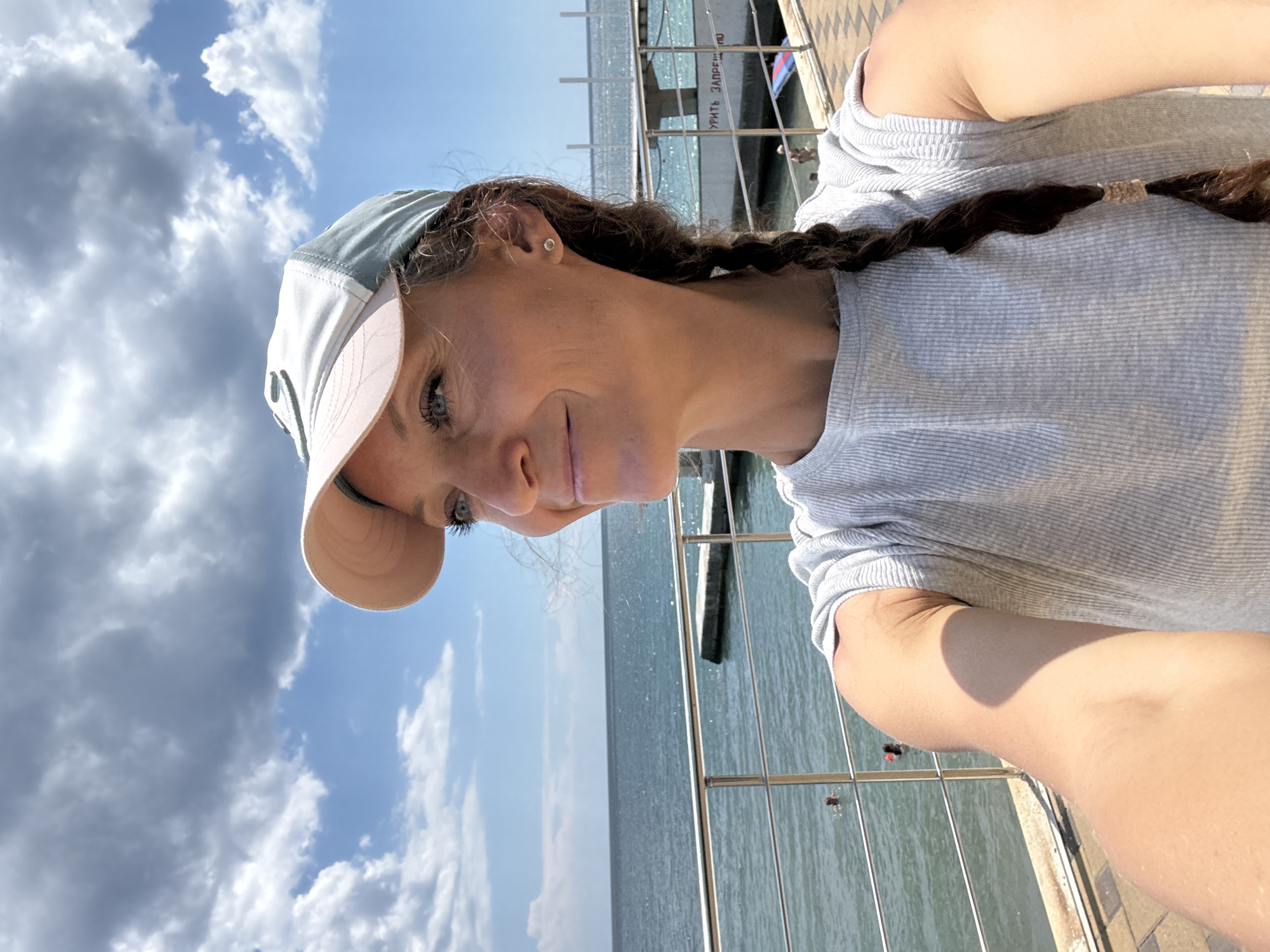 In Alushta, Ukraine, a woman with a braid of dark hair and a light-colored baseball cap is taking a selfie. She is wearing a grey t-shirt and stands on a tiled promenade overlooking the Black Sea. The sea is calm, with a few people swimming and a small pier visible. The sky is partly cloudy, with large, fluffy clouds against a bright blue expanse. The railing of the promenade separates the woman from the water, and a structure with white railings and text is visible further along the coastline. The sunlight casts shadows on the promenade and the woman's arm.