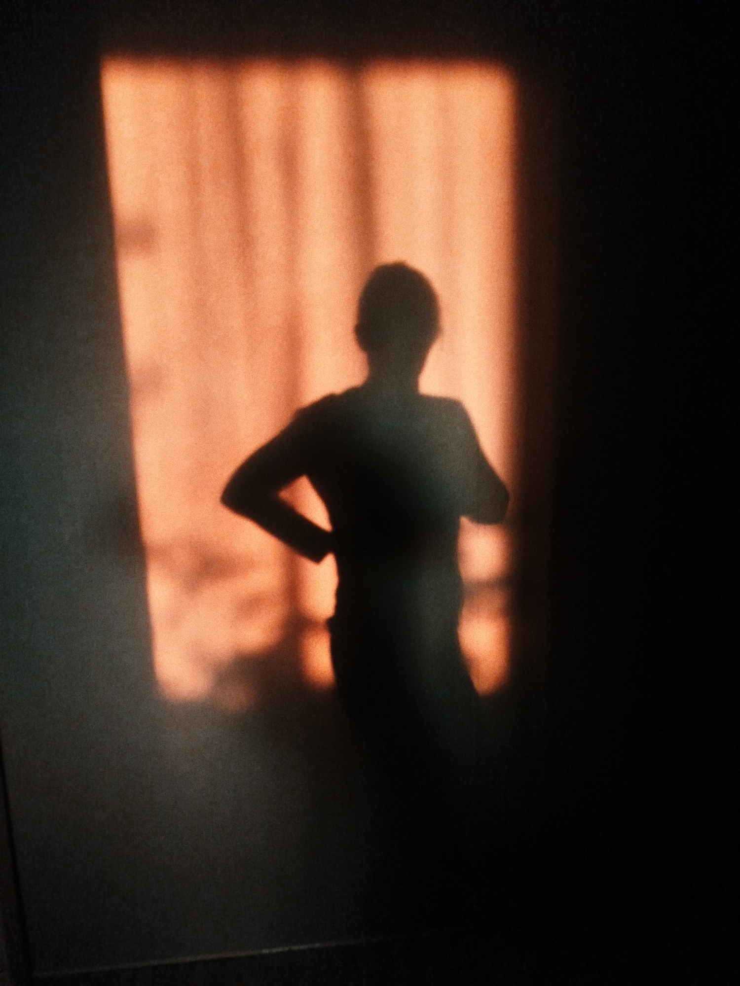 The scene depicts an interior space, likely a room, within the city of Izhevsk, Russia. A distinct human silhouette is projected onto a wall that is partially illuminated by a warm, orange-toned light. This light source, presumably a window, creates a pattern of vertical light and shadow stripes across the wall, indicating the presence of vertical blinds or a multi-paned window frame. The human figure's shadow shows an individual standing, with their left arm bent and hand resting on the hip/waist area. The right arm is also bent, held closer to the torso. No facial features, clothing details, or other specific objects are discernible beyond the shadow and light patterns. The immediate environment outside the illuminated area of the wall is dark, suggesting low ambient light.
