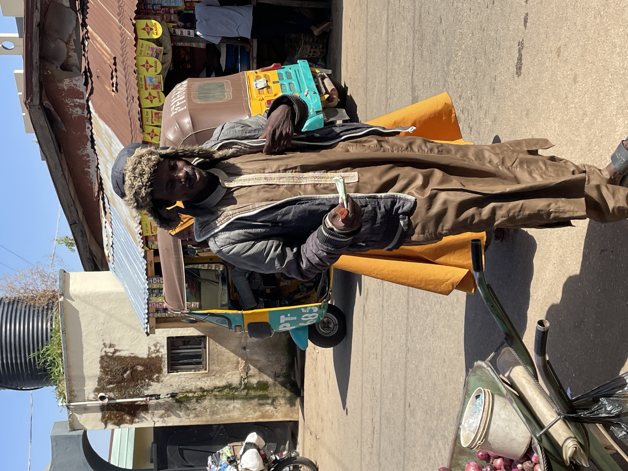 A man in Jos, Nigeria, stands in front of a yellow and turquoise auto rickshaw. He wears a fur-trimmed hat, a brown robe, and a gray jacket, holding a green bill in his hand. A roadside stall with "Maggi" signs is visible in the background. A wheelbarrow with onions and containers sits nearby.