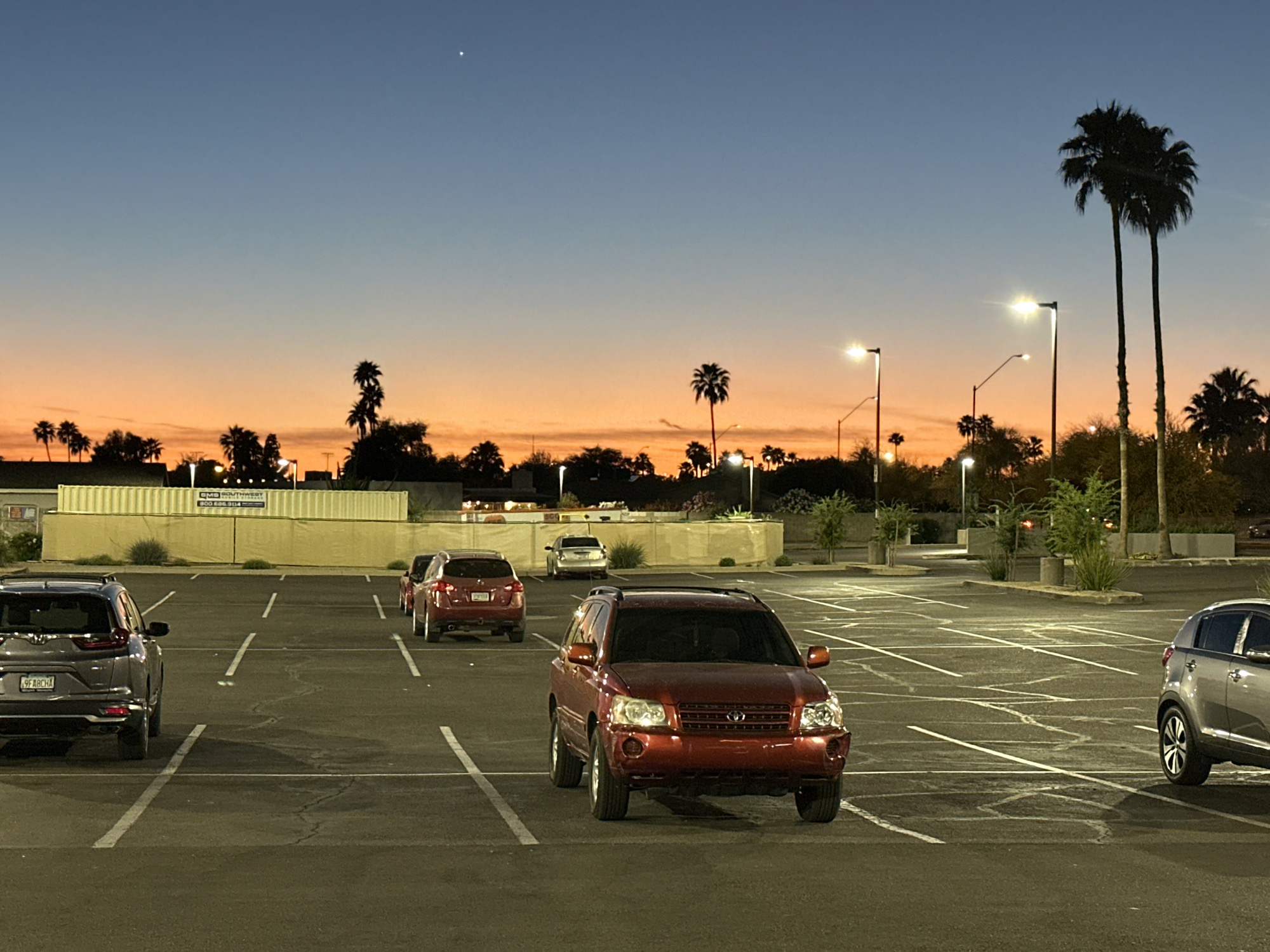 The image depicts an outdoor parking lot at dusk in Phoenix, Arizona, United States. The sky exhibits a gradient from deep blue at the top to a prominent orange and peach hue near the horizon, with a single bright celestial object visible in the upper left quadrant. Multiple artificial streetlights illuminate the asphalt parking surface, which shows white painted lines delineating parking spaces and some visible surface cracks.
Six vehicles are present:
1. A dark red SUV, possibly a Toyota Highlander, is positioned centrally in the foreground, facing towards the viewer and partially occupying a parking space.
2. The rear of a dark gray SUV, likely a Honda CR-V with license plate "9FABCHA," is visible on the far left, parked perpendicular to the camera.
3. The rear of a silver SUV, possibly a Kia Sportage, is visible on the far right, parked perpendicular to the camera.
4. The rear of a dark red sedan is parked in the mid-ground, left-center, facing away.
5. The rear of another dark red sedan is visible behind the first sedan, partially obscured.
6. The rear of a light-colored sedan is parked further back in the mid-ground, center-right.
Numerous palm trees and other silhouetted trees are visible across the horizon, characteristic of a desert environment. In the background on the left, a long, light-colored modular structure or container system is present, bearing a sign with text "CMG SOUTHWEST" and "800.685.9194" partially visible. No individuals are present, and no active interactions are depicted.