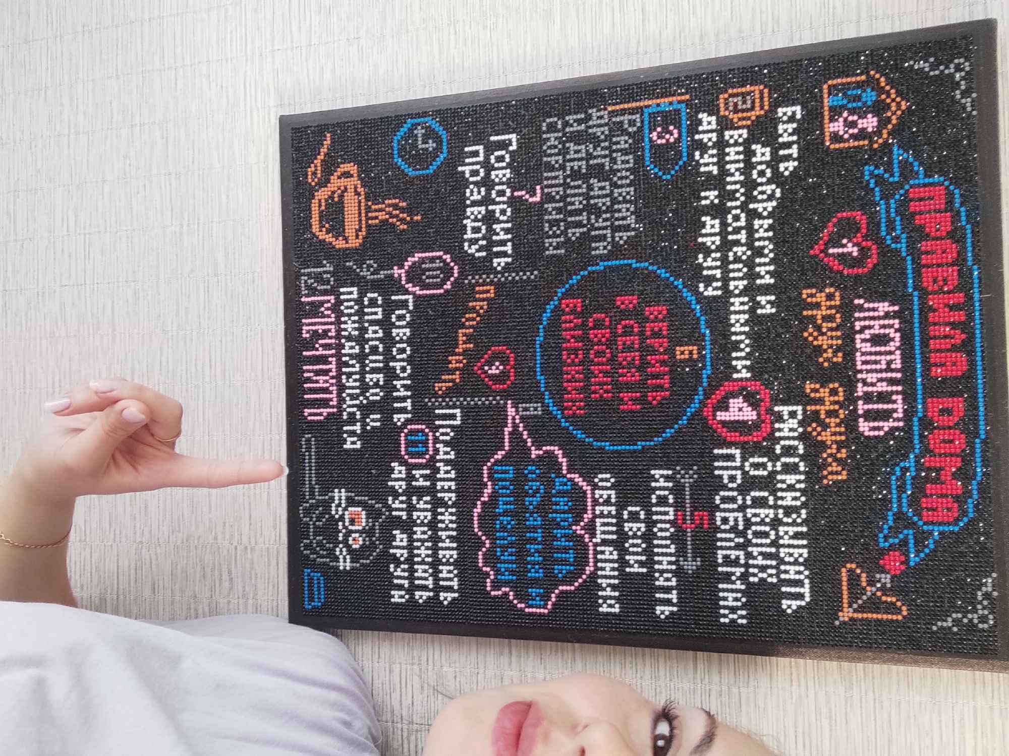 A woman is lying on her back, facing the camera and pointing with her right hand to a black-framed, rectangular board. The board is decorated with colorful, pixelated designs and text in Russian. The text appears to be a list of "rules for the house," including being kind, loving, telling the truth, and forgiving. The image is taken from a close-up perspective, focusing on the board and the woman's face and hand. The location of this photo is unknown, though the board suggests a domestic setting and the fact that the text is in Russian may indicate the city of Chaikovsky, Russia, as it is the location provided in the prompt.