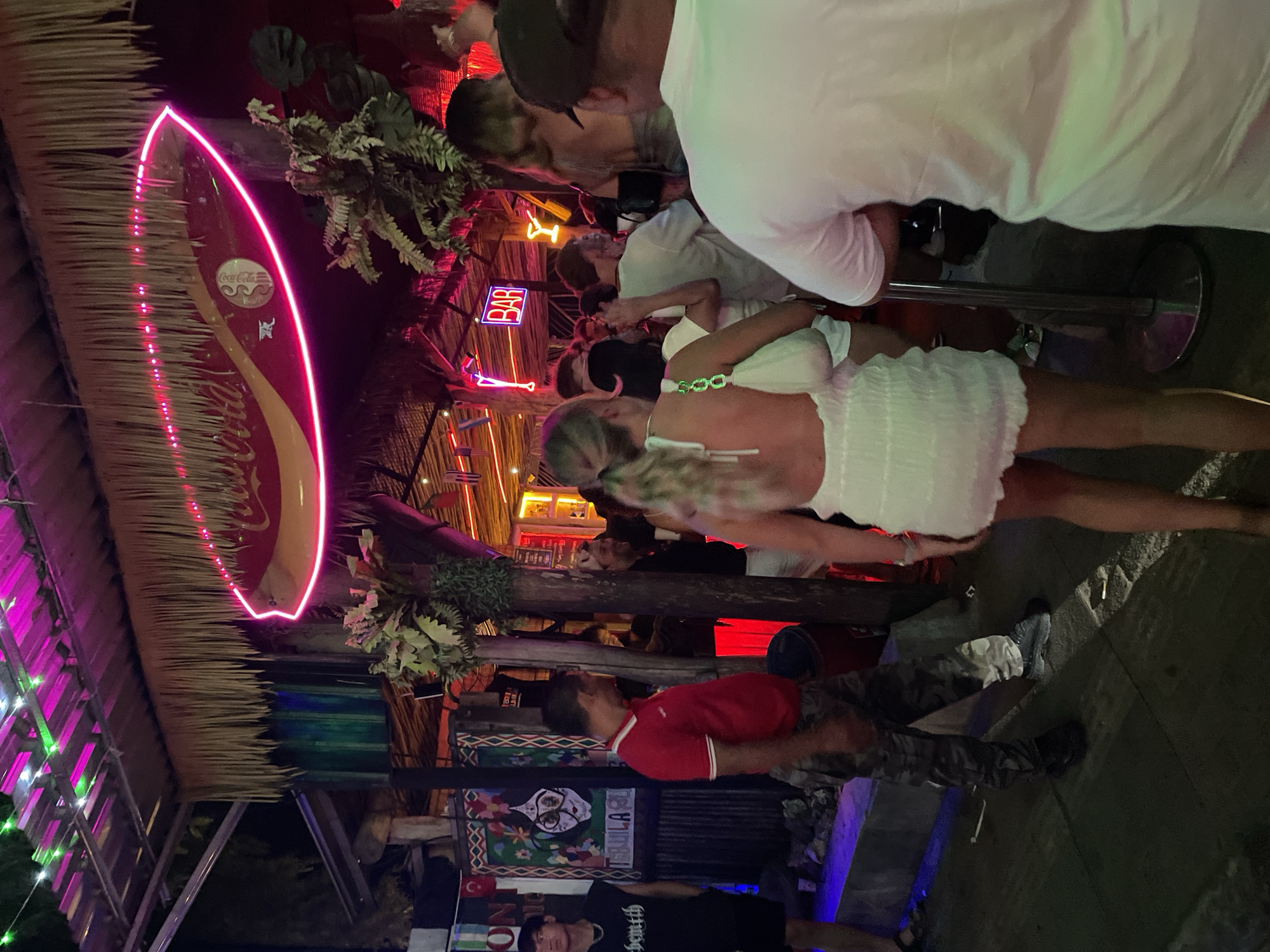 The image captures a lively outdoor scene at night, likely a bar or entertainment venue in an unknown city in Indonesia. The foreground features several people, with a woman in a white dress prominent on the right, her back to the camera. To her left, another woman in a white dress is also facing away, holding a white purse. A man in a red polo shirt and camouflage pants stands to the left of the women, facing towards the right. In the background, a crowd of people are visible, with neon lights illuminating the area. A prominent neon sign reading "BAR" is visible above a cluster of people. A large, surf-board-shaped sign with a Coca-Cola logo and pink neon trim hangs overhead, partially obscured by thatched roofing. The environment appears to be a tropical setting, with lush greenery and what looks like bamboo or wooden structures supporting the roof. The ground is paved, and the overall atmosphere suggests a vibrant nightlife scene.