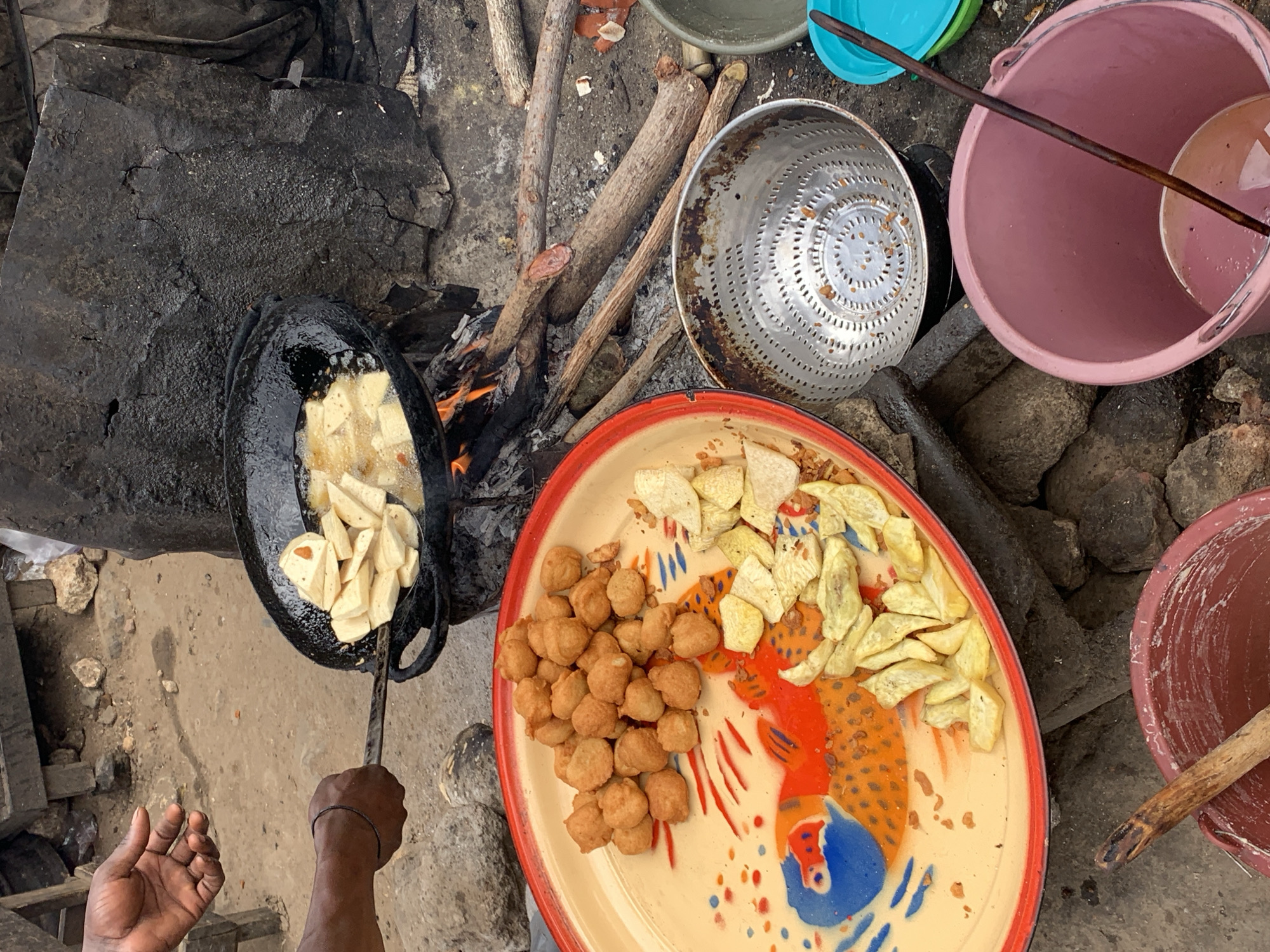 The image, taken from an overhead perspective, depicts a food preparation scene in Rafin Par, Nigeria. A dark-skinned individual is using a metal spatula to stir what appears to be diced plantains in a black pan of hot oil over a wood-fueled fire. To the right, a plate holds a pile of golden-brown, spherical fried dough and another pile of fried plantain slices. Adjacent to the plate are a metal colander and several buckets of various colors, indicating the possible separation of ingredients or use of multiple containers. The scene is set outdoors on a concrete surface among pieces of wood and rocks, suggesting an improvised cooking setup.