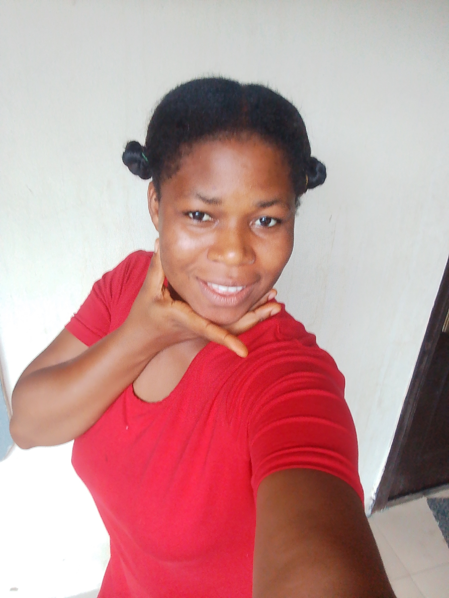 A young Black woman with dark skin and dark hair styled in two small buns is posing for a selfie indoors. She is wearing a vibrant red t-shirt and resting her chin on her hand, with her fingers extended across her cheek. She is smiling, displaying her teeth, and looking directly at the camera. The background appears to be a plain, light-colored wall, with a dark doorway visible on the right side of the frame. The lighting suggests an indoor setting, possibly a home or office in Yenagoa, Nigeria.