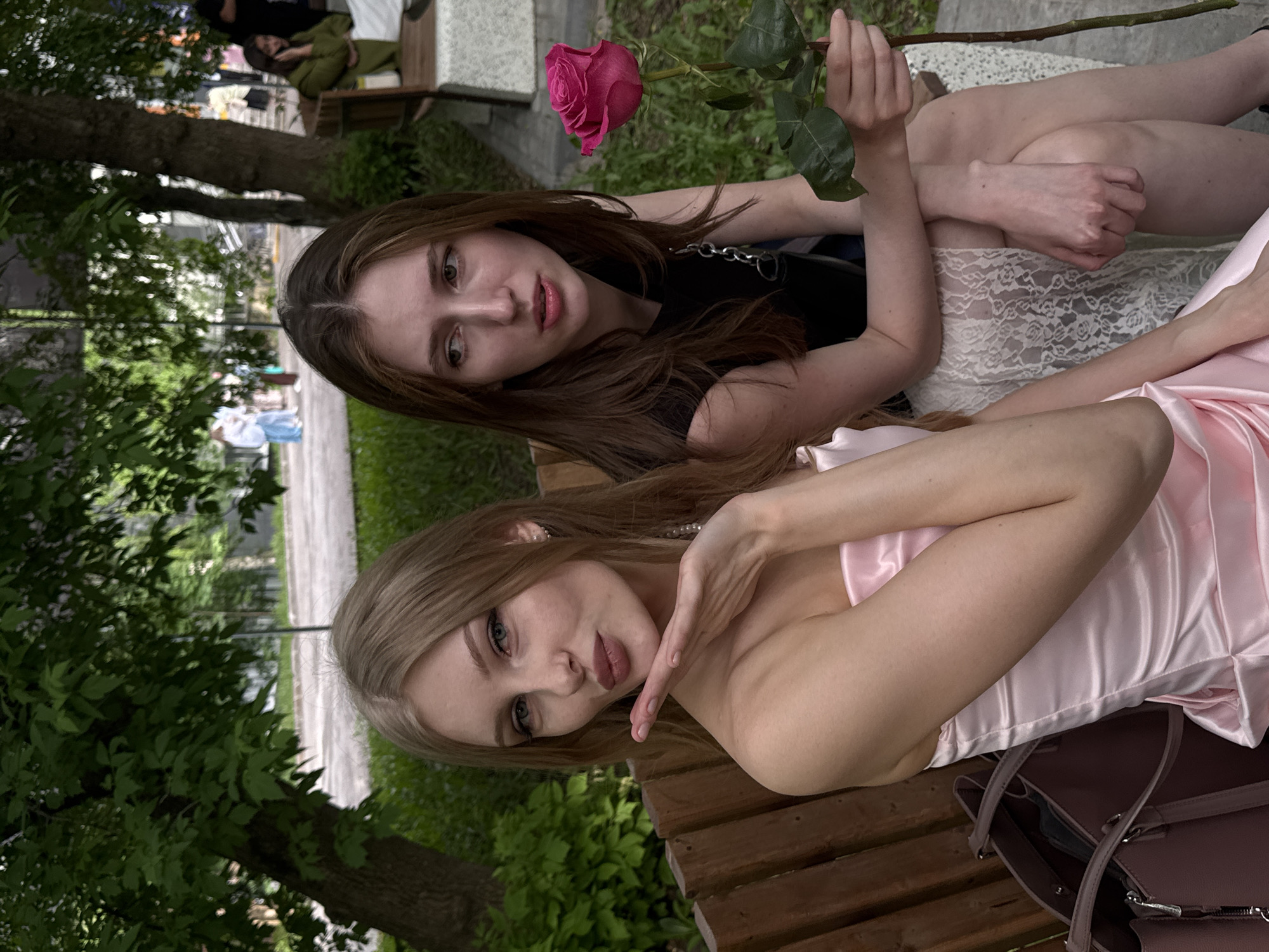 Two young women are sitting on a wooden bench in a park, likely in Perm, Russia, based on visual cues such as the style of street furniture and landscaping. The woman in the foreground has blonde hair, wears a pink dress, and poses with her hand near her face. The woman behind her has long brown hair, wears a black top with a silver chain, and holds a pink rose. They are on a bench with a brown leather bag underneath the pink-dressed woman. In the background, other people are seated on a bench, and greenery, trees, and a sidewalk suggest a public park setting.
