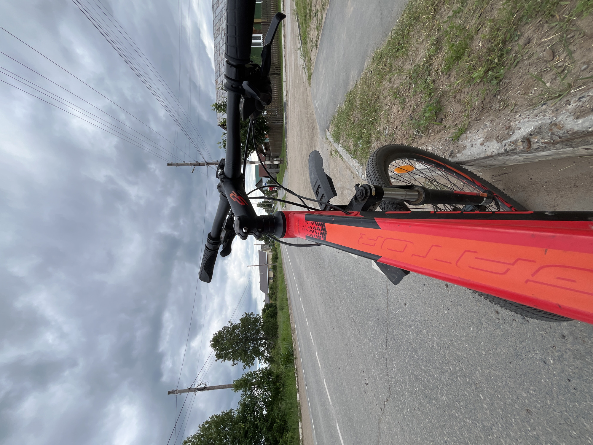 The image depicts a first-person perspective of someone riding a bicycle in Chaikovsky, Russia. The bicycle is bright red and black, with prominent branding visible on the frame. The scene is set along a paved road, with a dirt shoulder and grassy verge to the right. In the background, houses and trees line the roadside, suggesting a suburban or rural setting. Overhead, a network of power lines crisscrosses the sky, which is filled with heavy, dark clouds, indicating overcast and possibly impending rain. The time of day appears to be daytime, but the weather conditions contribute to a somber atmosphere. The road itself has white dashed lines marking the lanes. The focus is on the bicycle's handlebars and front wheel, with the road and surrounding environment receding into the distance. There is no visible text on the bicycle other than branding logos.