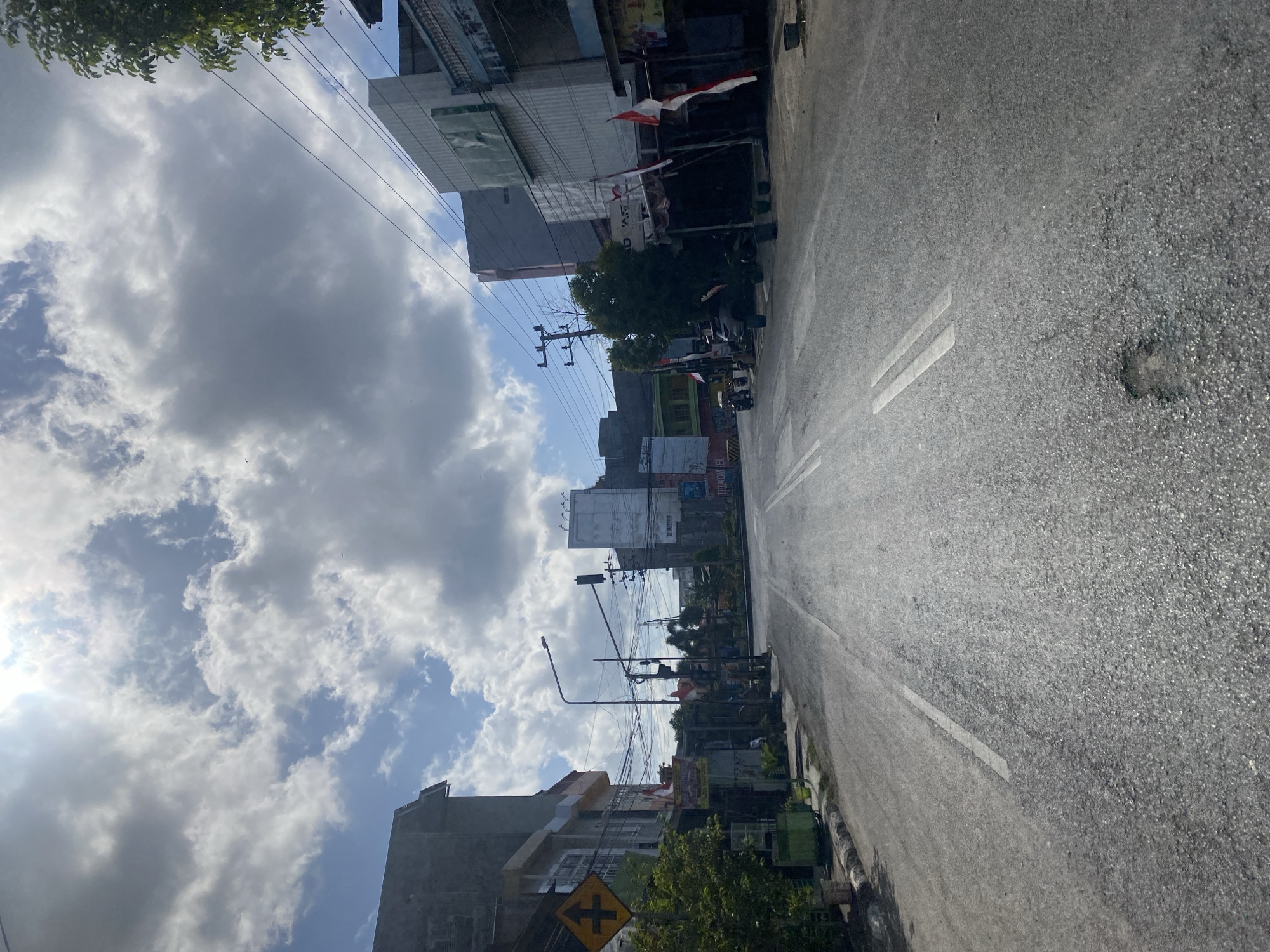 The image depicts a street scene in Bentengnge, Indonesia, under a partly cloudy sky with bright sunlight. The perspective is from the middle of the asphalt road, looking forward. On the left, a two-story building with a gray facade is visible, featuring windows on the upper level and a yellow traffic sign with a black symbol of four arrows pointing in opposing directions at the lower left. Adjacent to this building, a tree with green foliage grows. Further down the street on the left, several potted plants are arranged along a curb.
The road itself is marked with white lines, with a double white line in the center and a single white line to the right. There are visible patches of wear and a pothole on the road. Traffic lights are present, with a red light illuminated on one signal.
On the right side of the street, several buildings of varying heights and architectural styles line the road. One building has a light-colored facade with what appear to be advertisements or signage. Another building closer to the foreground has a dark awning supported by pillars, with Indonesian flags (red and white) displayed.
A few objects are visible on the street, including motorcycles, a car, and some trash cans. The overall impression is that of a relatively quiet street, possibly during midday or afternoon, with evidence of commercial activity and urban infrastructure. The sky is dynamic with large, white, and gray clouds, suggesting fluctuating weather conditions.