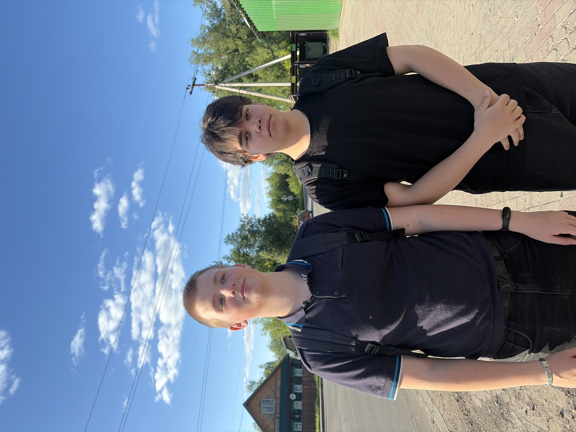 Two young males are depicted standing outdoors on a paved pathway adjacent to a dirt road under a clear blue sky with scattered white clouds, in what appears to be a semi-rural setting within the city of Semyonov, Russia. The sun is high, indicating midday or early afternoon.
The male on the left, with short light-brown hair and light complexion, wears a dark blue polo shirt with light blue and white striped accents on the collar and sleeves, dark jeans, and a black backpack. A silver-toned bracelet is on his left wrist and a dark fitness tracker on his right. He is looking upwards and slightly to his right with a subtle, content expression.
The male on the right, with dark, medium-length hair and light complexion, wears a plain black t-shirt, dark jeans, and a black backpack. His hands are clasped in front of his waist, and he maintains a neutral, direct gaze.
In the background, to the left, a traditional two-story wooden house with a dark roof and a fence is visible, surrounded by green trees and bushes. A light-colored vehicle is partially visible on the road in the mid-distance. To the right, a utility pole with power lines extends across the frame, and a green corrugated metal structure is present. The ground beneath the individuals transitions from a dirt shoulder to a light-colored paved pathway.