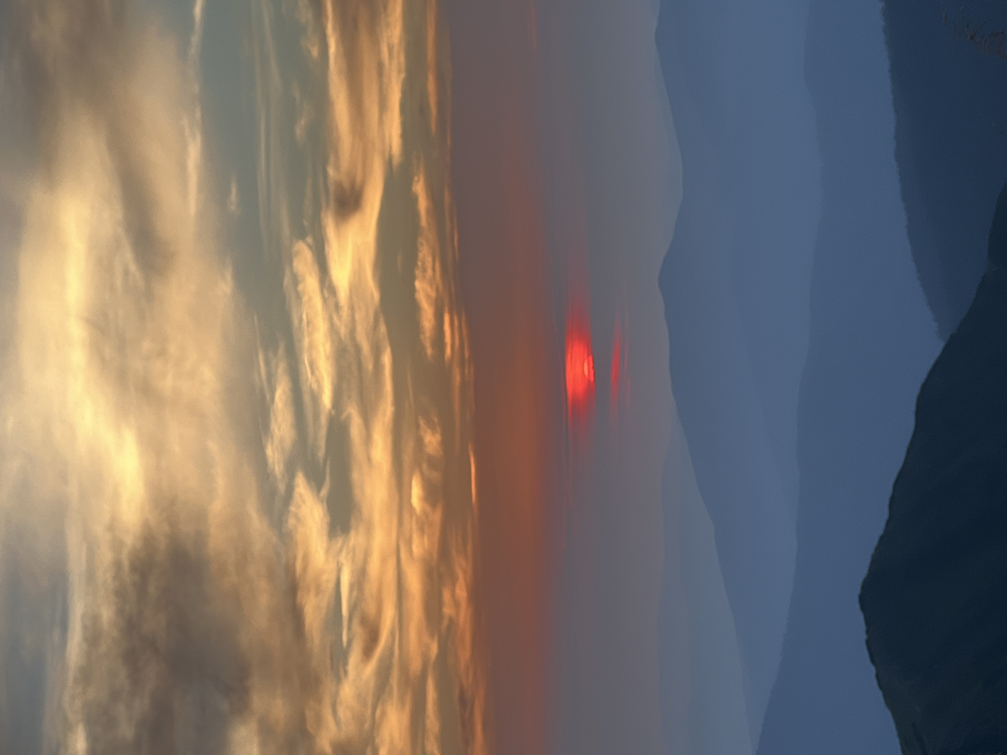 This is a vertical, high-angle, eye-level, medium shot of a mountain range at sunset. The sky is filled with orange, yellow, and gray clouds, with the sun appearing as a bright red disc near the horizon. The mountains are layered in shades of blue and gray, with the closest peaks appearing darker and more defined than those in the distance. The image captures the natural beauty of the Carpathian Mountains near the village of Luhy, Ukraine. There are no people or man-made objects visible in the scene. The environment is serene and majestic, showcasing the dramatic colors of the sunset over the rugged terrain.