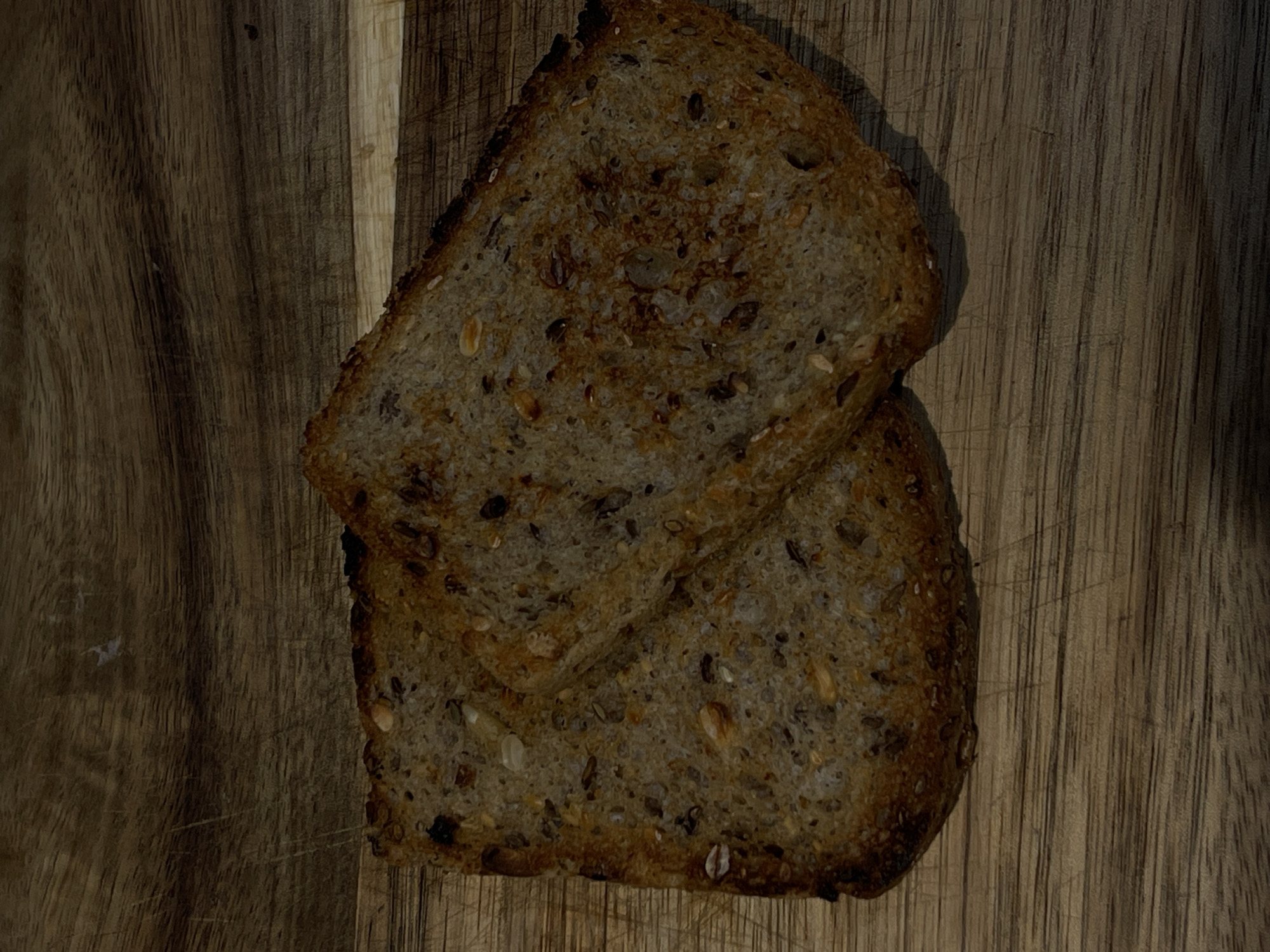 The image features two slices of dark multi-grain bread, partially overlapping, positioned on a wooden surface. The bread slices are rich in texture, with various seeds and grains like sunflower seeds, pumpkin seeds, and oats clearly visible throughout their composition. The crusts of the bread are dark brown, with some areas appearing more deeply toasted or slightly burnt. One slice is angled diagonally, resting atop the other.
The wooden surface below the bread displays prominent vertical wood grain patterns and subtle scratches, suggesting it might be a well-used cutting board or a rustic wooden table. The lighting is somewhat dim, creating soft shadows that add depth to the scene, particularly from the bread onto the wooden background. The light source appears to be from the upper left, accentuating the textures of both the bread and the wood.
There are no people or discernible activities taking place. The setting appears to be indoors. No specific time of day or weather conditions can be determined, and no text is visible within the image.