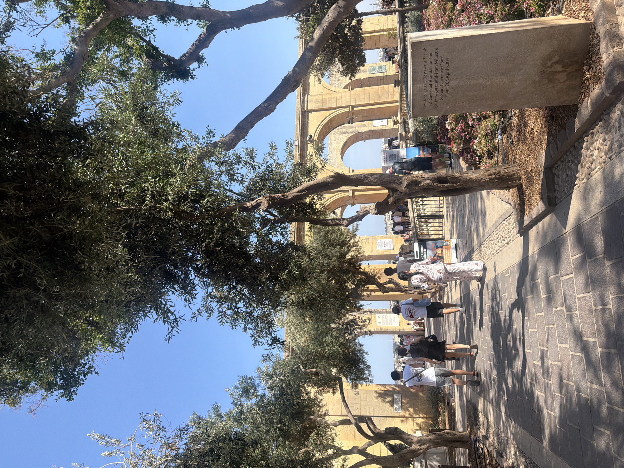 The image is a street scene in Valletta, Malta, featuring people walking along a stone-paved pathway lined with trees. An architectural structure with multiple arches stands prominently in the background. A stone plaque with inscriptions stands to the side of the path. Inscription mentions 'Barrakka'. Some people are standing or walking in groups near the arches and along the pathway. The sky is clear and blue, and sunlight filters through the trees, casting shadows on the ground.