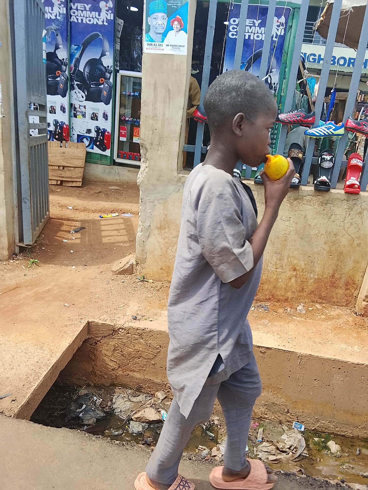 A young boy, appearing to be in his early to mid-childhood, walks along a dusty path in Akwanga, Nigeria. He is dressed in a light gray tunic over matching trousers and wears light-colored slip-on sandals. He holds a bright yellow, spherical fruit or object to his mouth, seemingly drinking or eating from it.
To his right, an open concrete drainage ditch, filled with stagnant water and various pieces of litter including plastic bags and wrappers, runs alongside the path. The ground is a reddish-brown dirt, uneven and with scattered debris.
In the background, a commercial area is visible. On the left, large blue and white posters for "COMMUNICATION" services, featuring headphones and other electronics, are displayed behind a partial metal gate. Text on a poster identifies "INGR. A.A. SULLE" and "DR. RAKSANA SUNDAY" as "MEMBER". Further right, behind another metal grate, various shoes and slippers, including colorful athletic footwear and patterned slides, are displayed for sale. A partial sign in the background reads "NIGHBOR" (likely "Neighbor"). Other storefront details and indistinct text like "40" and "THEOC" are visible. The scene is brightly lit, indicating it is daytime.