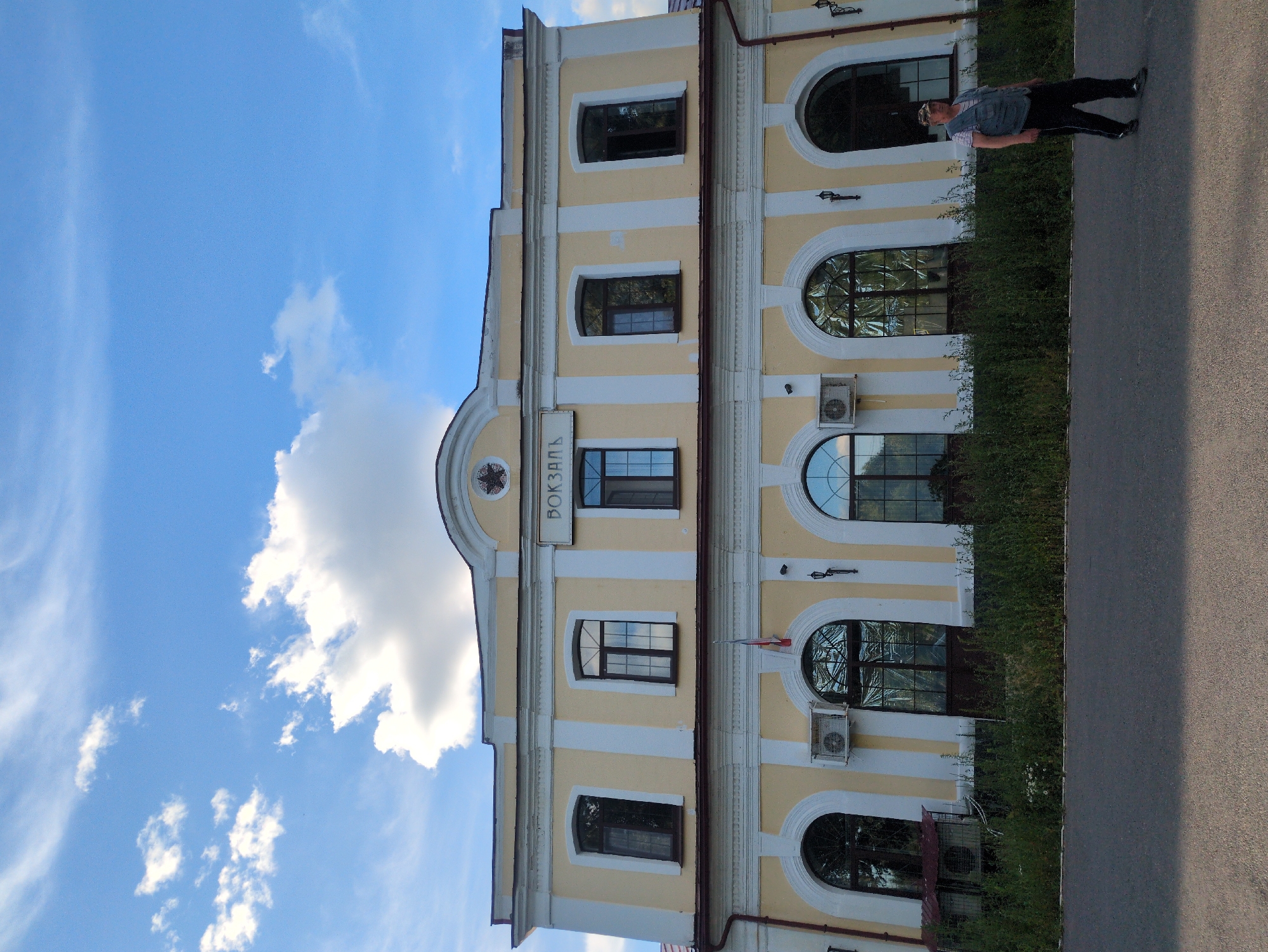 The image depicts a person standing in front of a light yellow, two-story building with white trim. The building has several arched windows with dark frames, and some windows are reflecting the blue sky and white clouds. There are also air conditioning units mounted on the building. A sign with Cyrillic text is visible above one of the windows. The building is flanked by a dense green hedge on one side and an asphalt path or road on the other. The sky is bright blue with scattered white clouds, suggesting it is daytime and the weather is clear. The person is wearing a blue vest over a striped shirt and dark pants, and appears to be looking towards the building. The overall scene has a slightly tilted perspective. The text on the sign appears to be "ВОКЗАЛ", which translates to "Station" in Russian, indicating this is likely a train station.