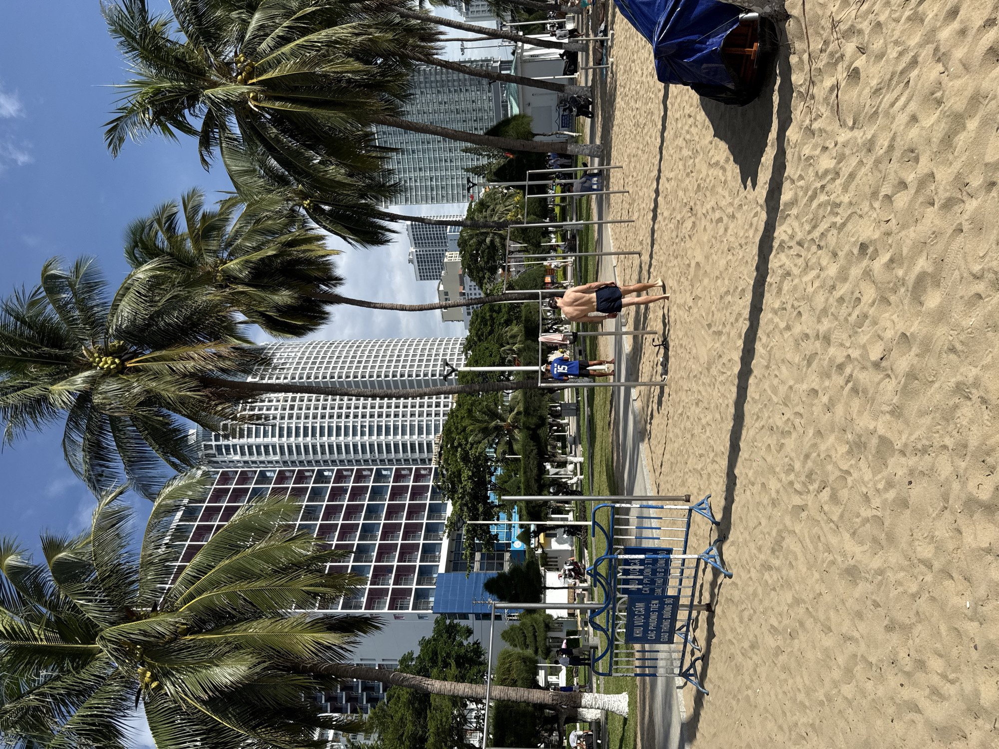 In Nha Trang, Vietnam, a sandy beach area features a makeshift outdoor gym with pull-up bars. Palm trees line the background, with tall, modern buildings rising behind them, suggesting an urban coastal setting. Two individuals are visible: one man, wearing blue shorts and a blue jersey with the number 15, is standing near the gym equipment, while another man, wearing dark shorts, walks away from the viewer towards the gym. A blue and white barrier, possibly indicating a restricted area and displaying text in Vietnamese, is positioned in the foreground. The sky is clear with some scattered clouds.