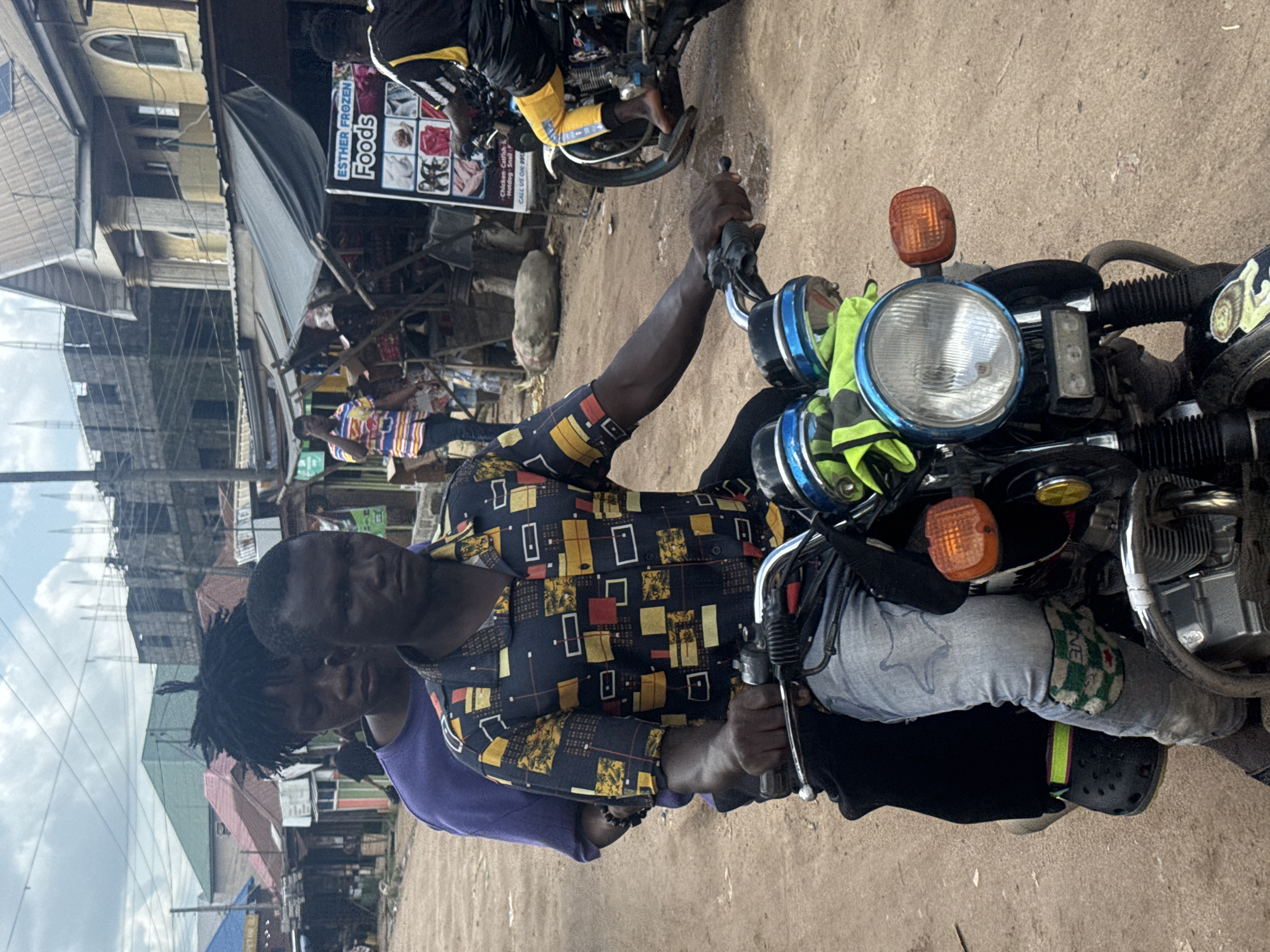 Two men are on a motorcycle in an outdoor market setting. The man in the foreground is wearing a colorful, patterned shirt with black, yellow, and red geometric shapes, and a purple t-shirt underneath. He has dreadlocks and is looking towards the camera. The second man, partially visible behind him, is wearing a black shirt with yellow accents. The motorcycle is black with a prominent round headlight and an orange turn signal. A neon green cloth is draped over the handlebars. In the background, there are market stalls with various goods, including food items displayed on a sign that reads "ESTHER FROZEN FOODS". The ground is a dusty, sandy surface typical of a market. The scene appears to be in daylight, with a mix of sunlight and clouds in the sky. The overall impression is a bustling marketplace in Ihiagwa, Nigeria.