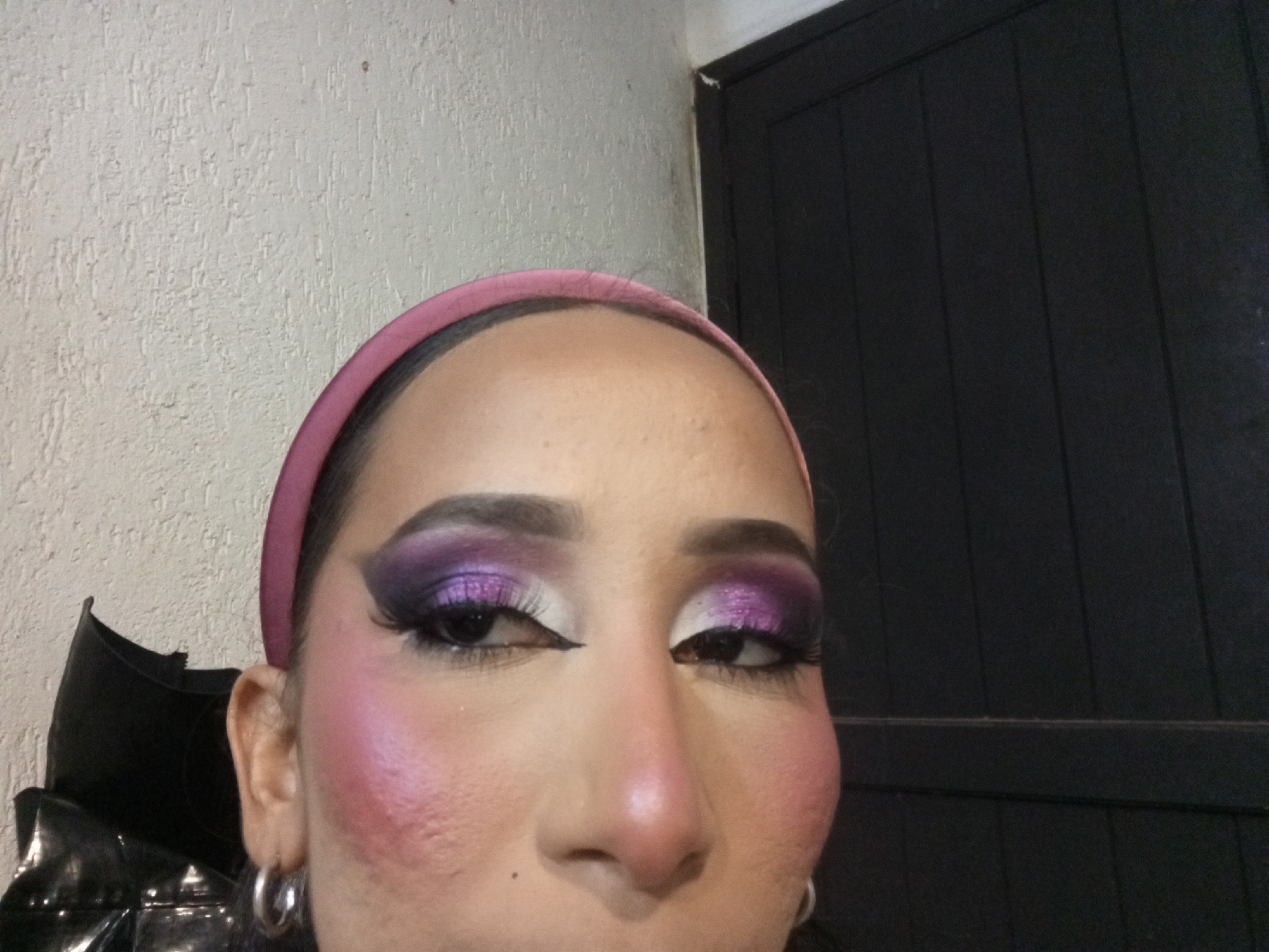 A close-up photograph shows a person with fair skin wearing a pink headband and elaborate purple and white eye makeup. The person has dark eyebrows, long eyelashes (likely artificial), and pink blush on their cheeks. The person is wearing silver hoop earrings. A black shiny object is visible in the lower-left corner. The background consists of a textured light-colored wall and a dark, possibly black, wooden door with vertical panels. The location of the photograph is not evident from the image. Without additional information, it is impossible to confirm whether this photo was taken in Porlamar, Venezuela.