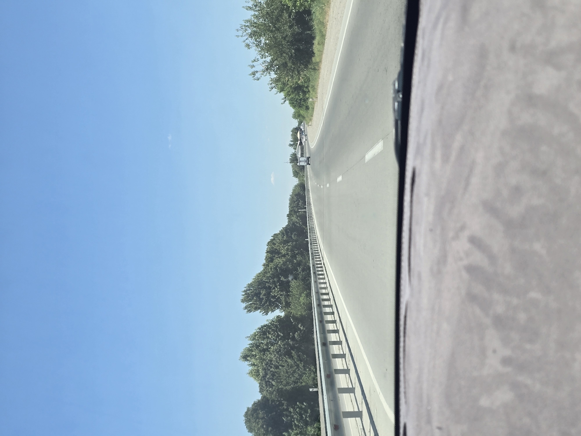 The image shows a view from inside a vehicle traveling on a highway. The highway is paved with asphalt and has white lane markings. A metal guardrail runs along the left side of the road. On the left side of the highway, there is a line of trees. On the right side of the highway, there are also trees and some vegetation. The sky is clear and blue. In the distance, there are a few vehicles on the road, including a white truck and a white car. The foreground of the image shows the dashboard of the vehicle, which is gray and textured. This scene is consistent with a highway in the city of Rabatiohun, Uzbekistan, under a clear sky during the day.