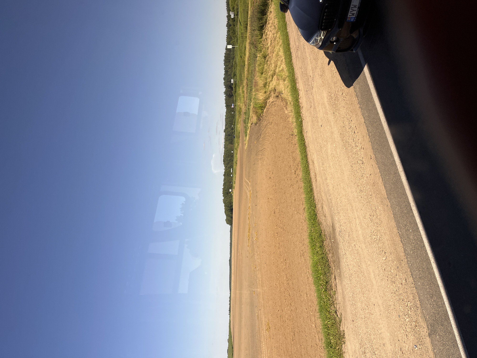 The image shows a view from inside a vehicle, looking out at a rural landscape. The foreground features a dirt road with a black car parked on the right side. Beyond the road is a partially plowed field with brown soil, transitioning to green grass along the edge. In the distance, a line of trees forms a dark green border against a clear blue sky. Reflections of the vehicle's interior are visible on the window. The scene appears to be in the vicinity of Юнькаўскі сельскі Савет, Belarus.