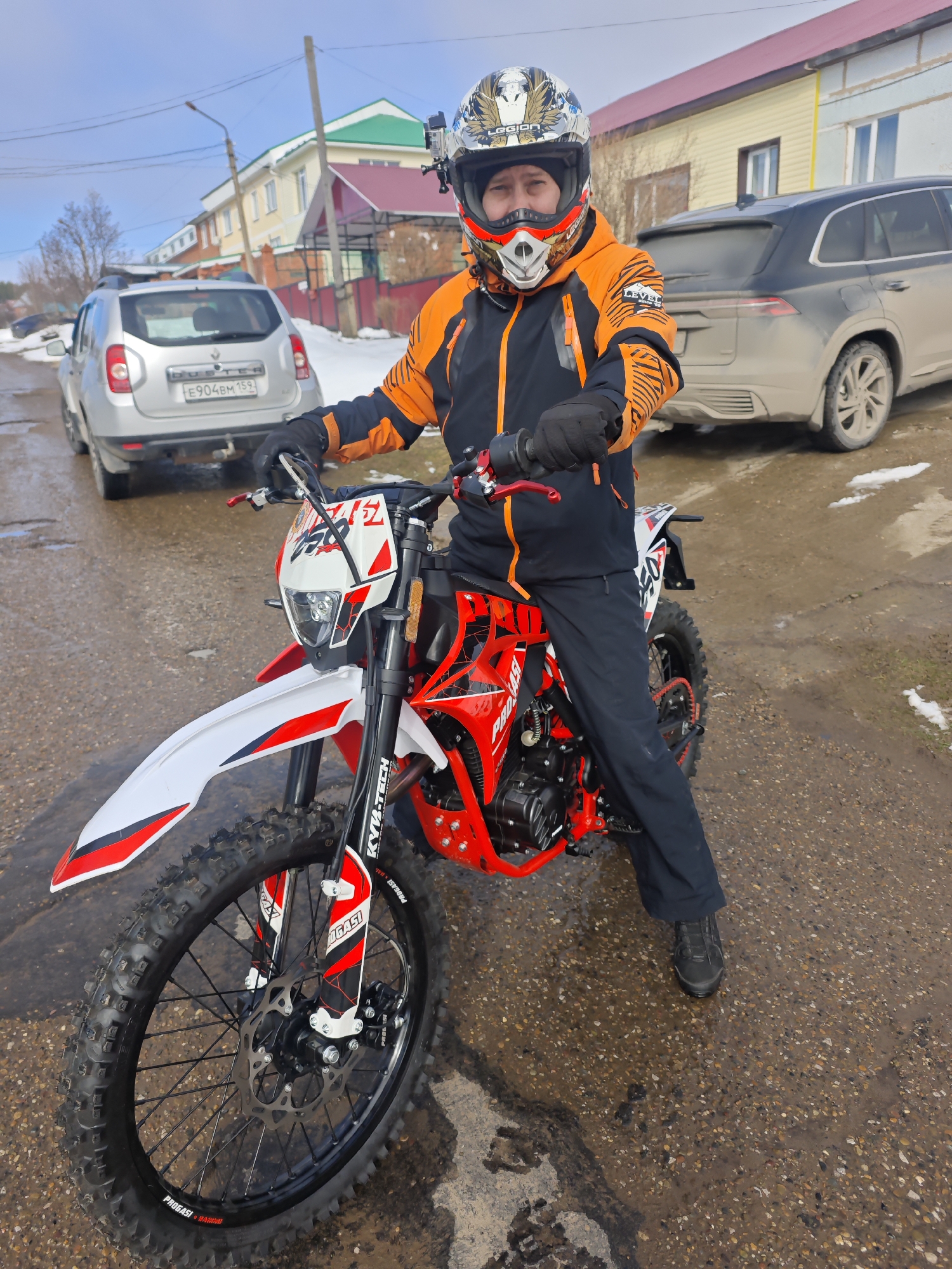 A man wearing a helmet and an orange and black jacket is sitting on a red and white dirt bike on a paved road. He is wearing black gloves and pants with black shoes. The bike has the words "PROGASI" on the frame. A silver Renault Duster with the license plate "E904BM159" is parked behind the bike on the left. A dark gray SUV is parked to the right of the bike. Houses and a street light are visible in the background under a partly cloudy sky. Patches of snow are scattered around. The location is Прикамский, Russia. The road appears wet.
