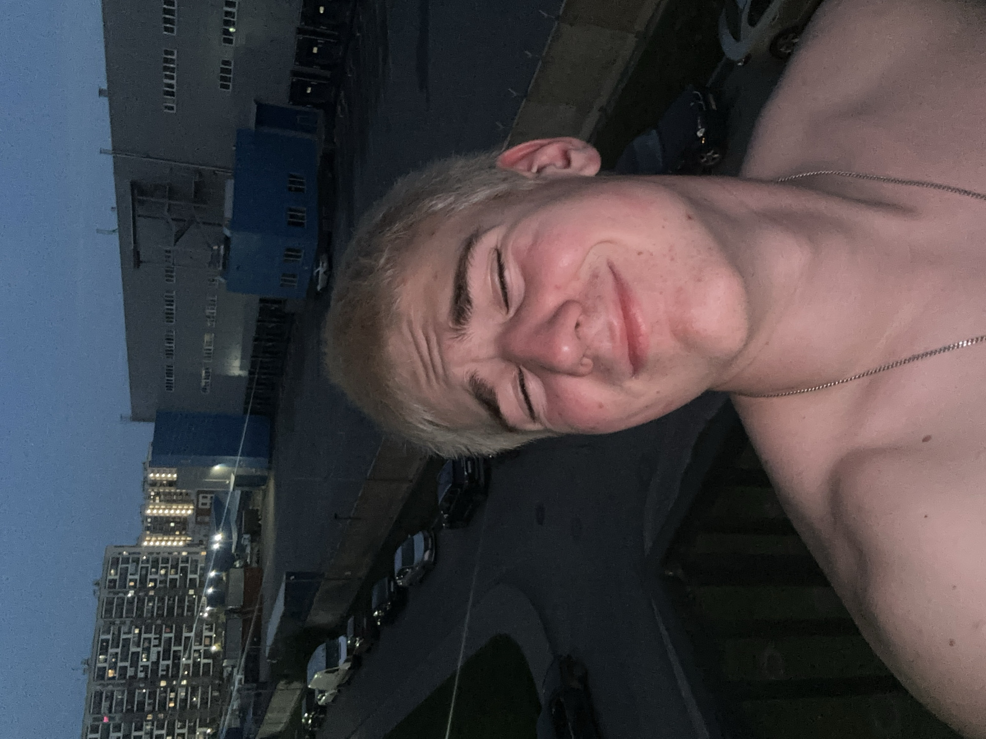 A casual selfie, rotated 90 degrees clockwise, captures a young, light-haired individual, likely male, at dusk or night. The subject, in the foreground, has fair skin and light blonde hair. He is shirtless and wears a thin, silver-colored chain necklace. His eyes are squinted or closed, and a gentle smile or playful expression adorns his face, creating wrinkles around his eyes and forehead, and giving his cheeks a rosy appearance.
The background reveals an urban or industrial environment. On the left, a large, multi-story grey building with numerous rectangular windows, some illuminated, stands against a dark blue-grey sky. A smaller, blue rectangular structure is positioned closer to this building. Further back and to the lower left, another tall building, possibly residential or commercial, is visible with many lit windows, reinforcing the nighttime setting. A dark asphalt road with parked cars runs between the foreground and the distant structures, suggesting the photo was taken from an elevated viewpoint overlooking the street. No text is visible in the image.