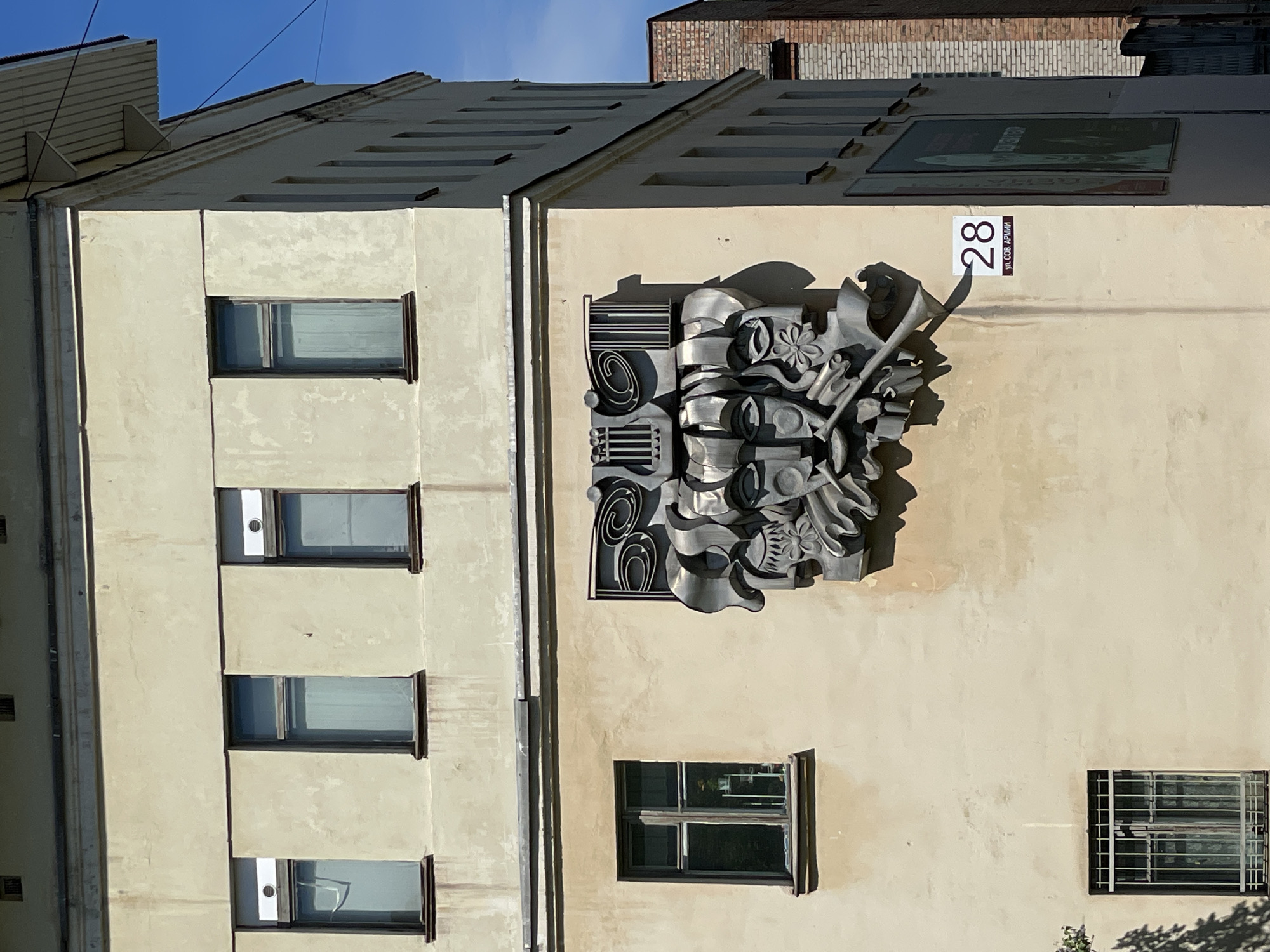 The image depicts the exterior facade of a multi-story building located at 28 Soviet Army Street (ул. Советской Армии, 28) in Zheleznogorsk, Russia. The building is constructed with light-colored, possibly stucco, walls in a pale yellow or beige hue, showing some signs of wear and staining.
On an upper floor, three rectangular windows with dark frames are visible. The left and rightmost of these windows have small, horizontal white ventilation units mounted above their frames. Below this level, a prominent horizontal band spans the facade.
Centered on the building's facade, between the upper and lower window levels, is a large, intricate metallic bas-relief sculpture. The sculpture is rendered in shades of grey and silver and features multiple stylized, abstract faces, a prominent lyre element at the top, and a horn or trumpet-like instrument extending from the lower right portion. It includes swirling, organic, and geometric forms. Direct sunlight from the upper right casts distinct, elongated shadows from the sculpture onto the building's surface.
Below the sculpture and to the left, a single rectangular window with dark frames is secured by a multi-paneled metal security grid or bars. Partial foliage from a tree is visible in the lower-left corner, casting shadows on the wall.
To the right of the sculpture, a white rectangular street sign with a dark border displays the number "28" in black at the top, and "ул. СОВ. АРМИИ" in smaller black text on a dark red-brown bar below it, indicating the street address.
The right side of the building is also visible, extending into the frame, revealing additional windows with dark frames and a slightly overhanging roof structure with exposed rafters. Further in the background on the right, a multi-story red brick building is partially visible. The sky above is clear and blue. No individuals are present in the scene.