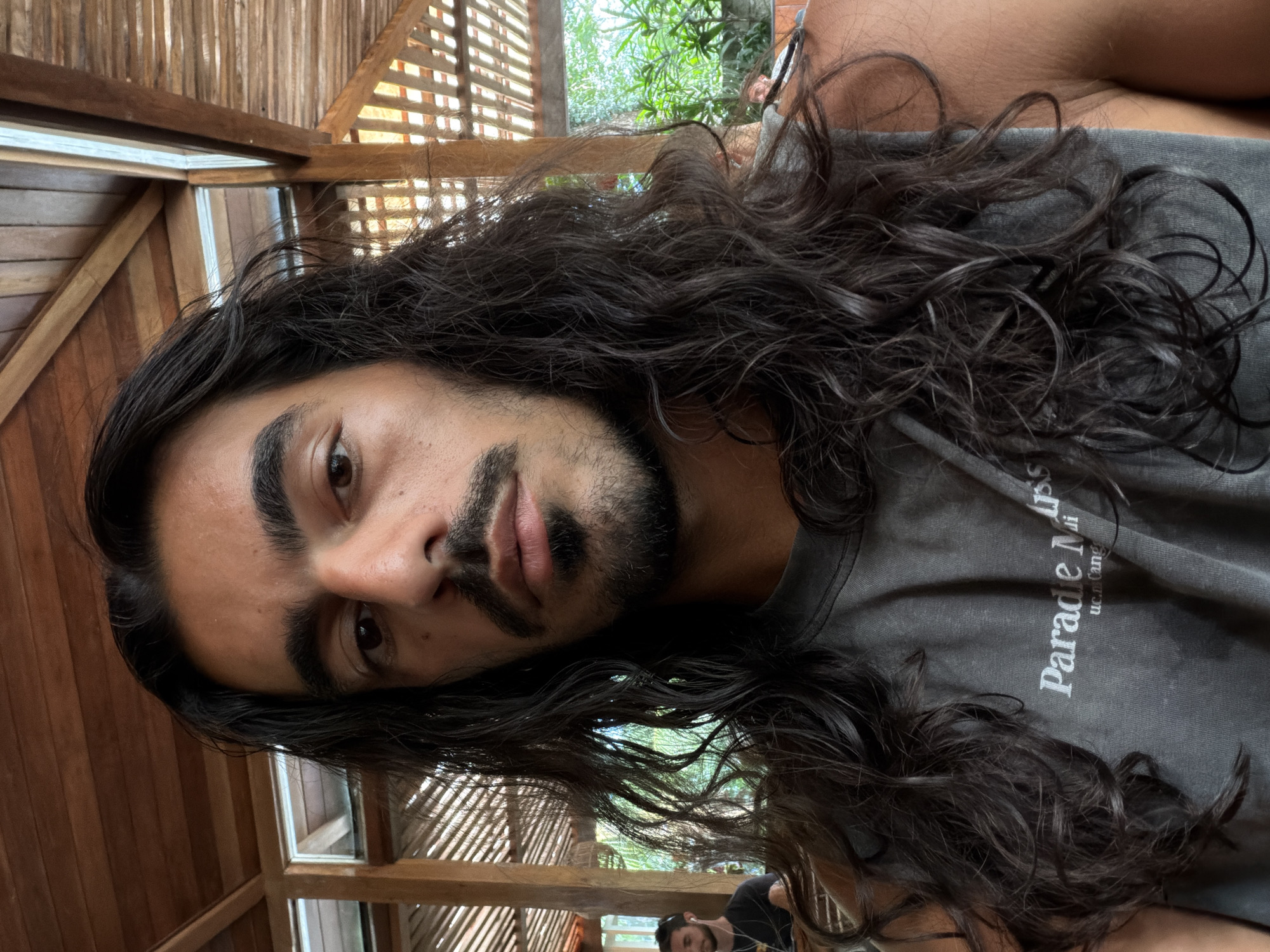A close-up, rotated portrait depicts a man with long, dark, wavy hair, dark eyes, and a dark mustache and beard. He is wearing a gray t-shirt with white text, the start of which reads "Parade". The setting appears to be a wooden structure with open latticework, possibly a gazebo or covered patio. In the background, greenery suggests an outdoor location. Another person with a beard and wearing a black t-shirt can be seen at the bottom of the frame. The location, Kuta Selatan, Indonesia, is provided as information associated with the scene.