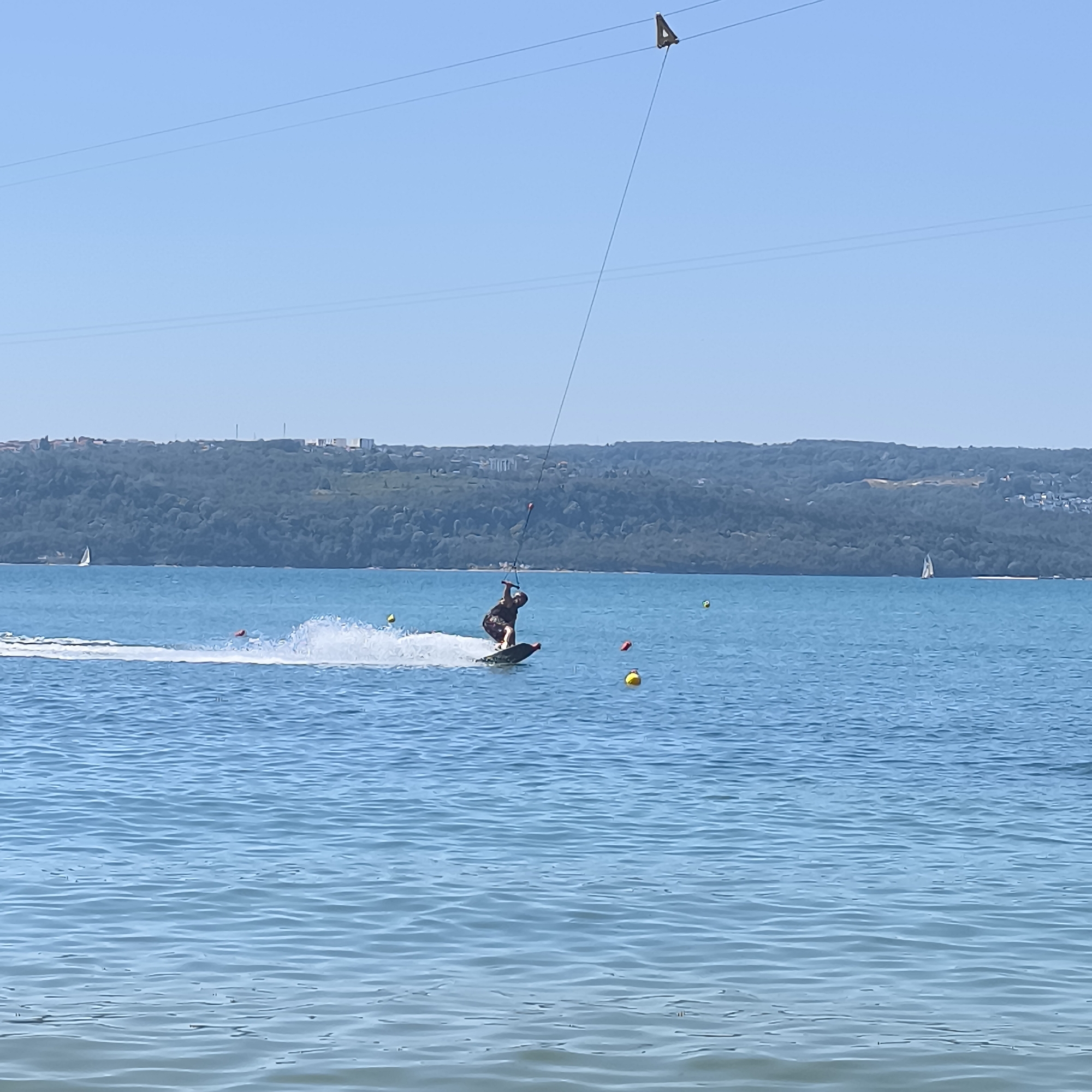 This is a photograph of a person wakeboarding on the water, likely in the Varna Bay area of Varna, Bulgaria. The sky is clear and blue, with power lines visible in the upper portion of the image, and a triangular marker hanging from one of the lines. The water is calm with some gentle ripples. The wakeboarder is in motion, creating a spray of water behind them. The wakeboard itself appears to be dark colored. In the background, there is a lush, green, forested hillside with some buildings visible on top, indicating a coastal city. There are several buoys floating in the water, one yellow and two red. A sailboat with a white sail is visible in the distance on the left side of the frame. The sun appears to be high in the sky, casting a bright light on the scene.