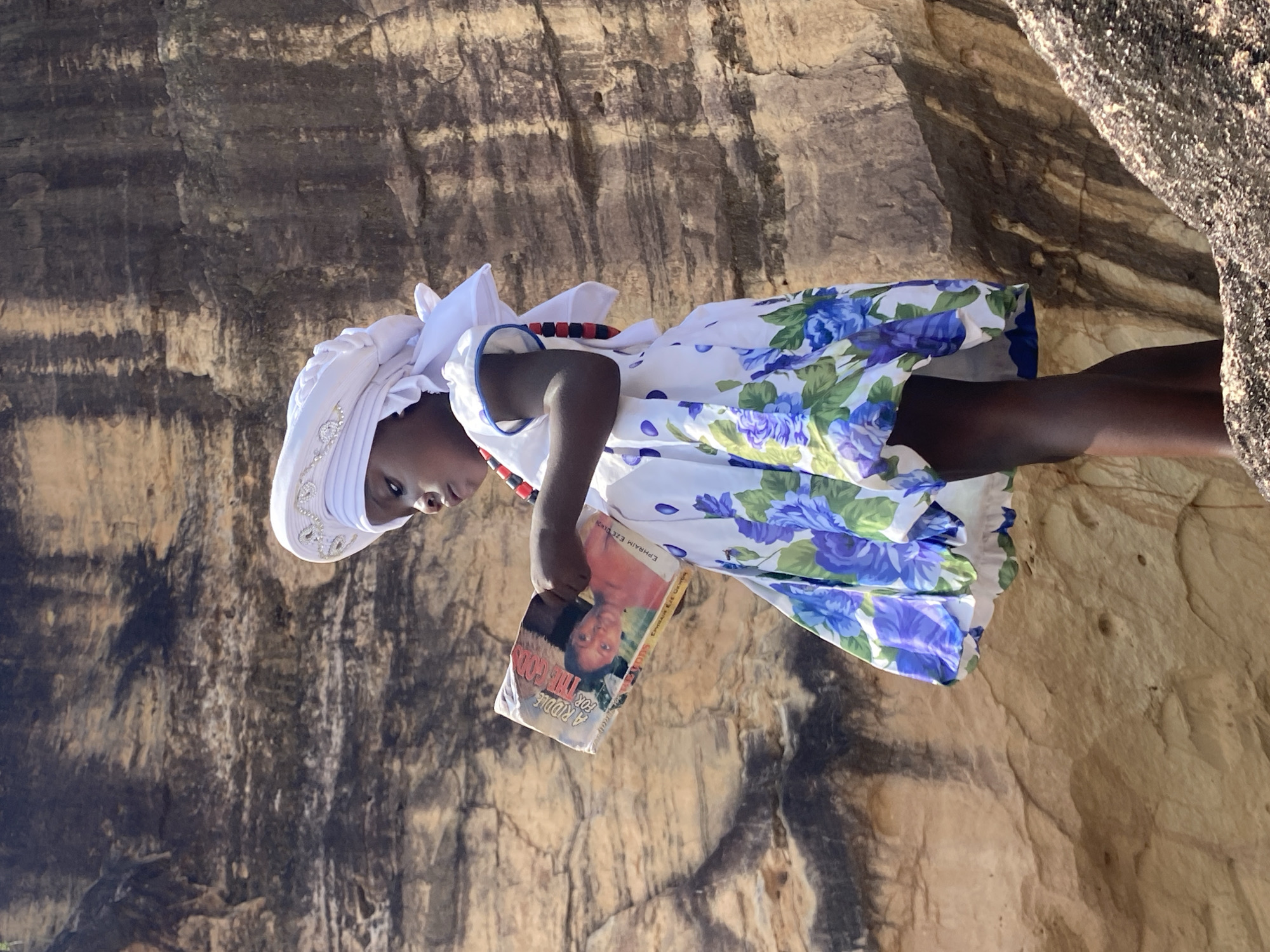 A girl is holding a book in front of a rock face in Amaseri, Nigeria. The girl is wearing a white head covering decorated with white rhinestones, a white, sleeveless dress patterned with blue and green floral designs, and a red and black beaded necklace. The book is titled "A Riddle For The Gods" by Ephraim Ezemba. The rock face behind the girl has alternating sections of dark and light rock.