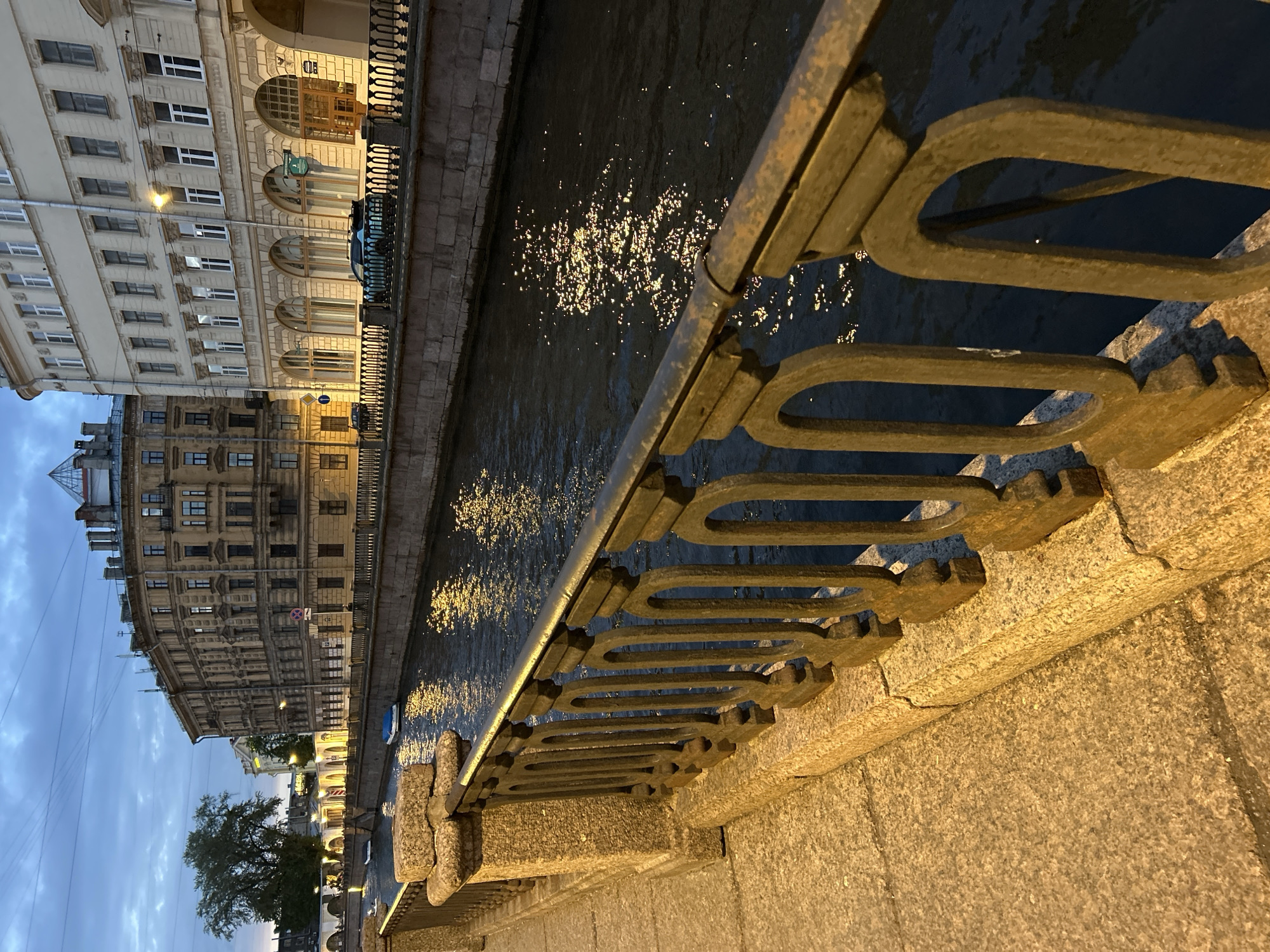 This is a photograph taken at dusk, looking out over a canal in an urban environment. In the foreground, a stone embankment with a decorative metal railing lines the edge of a canal. The railing is made of dark metal with an ornate, repeating oval design and is mounted on granite posts. The canal water is dark and reflects the lights from the buildings and streetlamps, creating shimmering patterns on its surface.
Along the opposite bank of the canal, several multi-story buildings are visible. These buildings appear to be of historical architecture, with classical facades, arched windows, and decorative stonework. The buildings are lit from within and by streetlights, casting a warm glow. A few small boats are moored in the canal, some covered with blue tarps.
In the background, more buildings and trees can be seen, receding into the dimming light of the twilight sky. The sky is a deep blue with hints of lighter clouds. The overall impression is of a serene and picturesque urban scene at the end of the day. The location is identified as Unknown, Unknown.