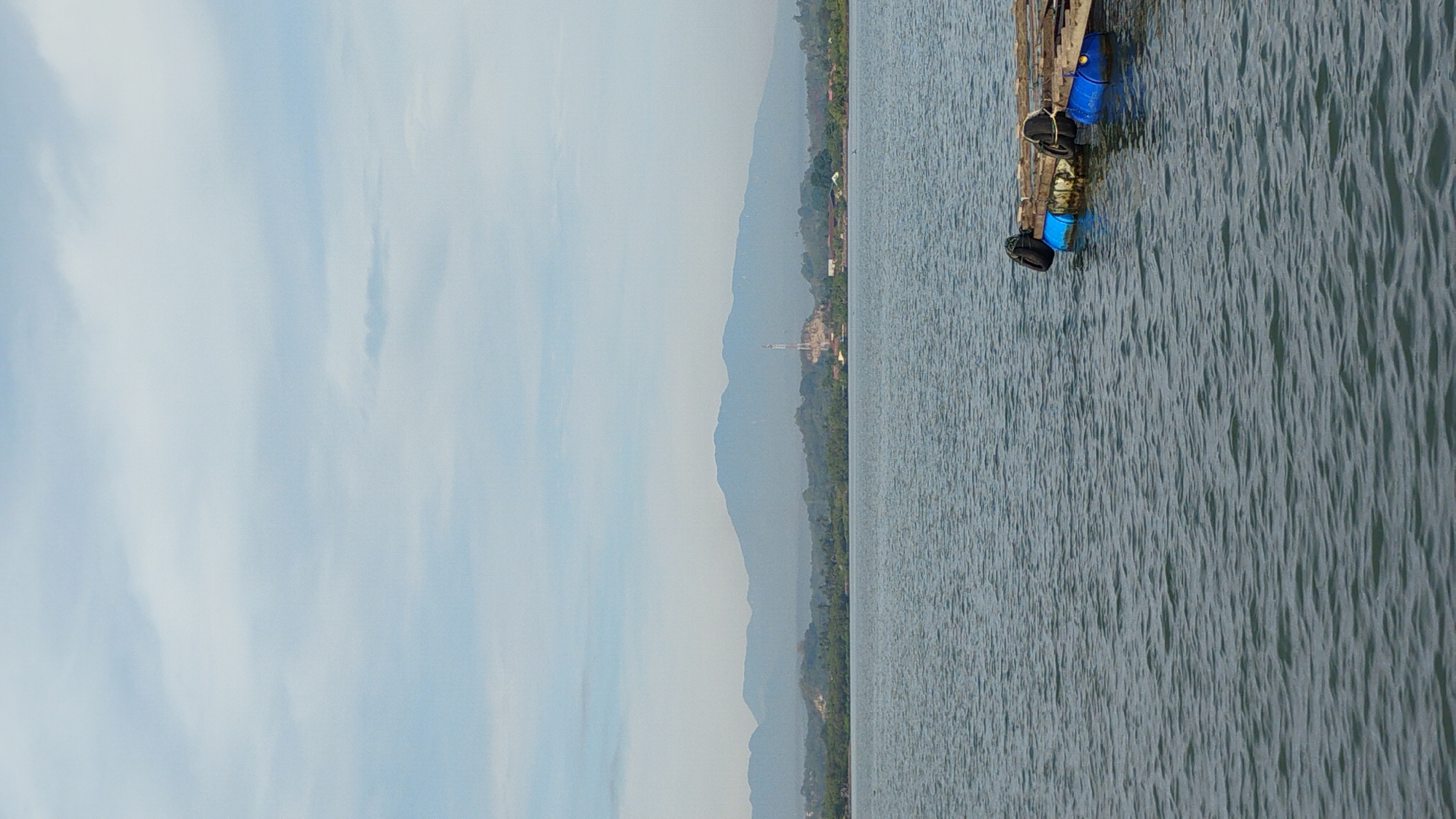 This image depicts a serene body of water with a distant shoreline featuring lush green trees and a mountain range under a cloudy sky. In the foreground, a wooden raft with blue barrels and black fenders floats on the water. The scene is likely the reservoir of the Crueng Mane dam in Lhokseumawe, Aceh, Indonesia, a significant water source for irrigation and a potential recreational area. No people are visible in the image.