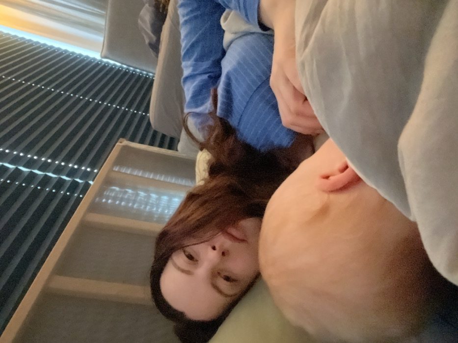 An adult female and an infant are situated in an indoor setting, identified as a private residence in Obermoschel, Germany. The female is positioned on her back, resting on a light-colored pillow, her head turned slightly towards the viewer. She has dark, wavy hair and is wearing a blue, long-sleeved, ribbed top. Her eyes are open, and her expression is neutral. Adjacent to the female, an infant lies, with only the head and a partial ear visible. The infant has light-colored hair and is positioned facing away from the viewer, largely obscured by a light grey or light blue duvet or blanket. The female's right arm is extended across her midsection, partially under the blanket. In the background, a light-colored headboard is visible, along with horizontal blinds covering a window, through which light is filtering, creating a striped pattern. A light-colored sofa or chair is partially visible in the upper right quadrant of the image. The scene appears to be a resting moment within a domestic environment.