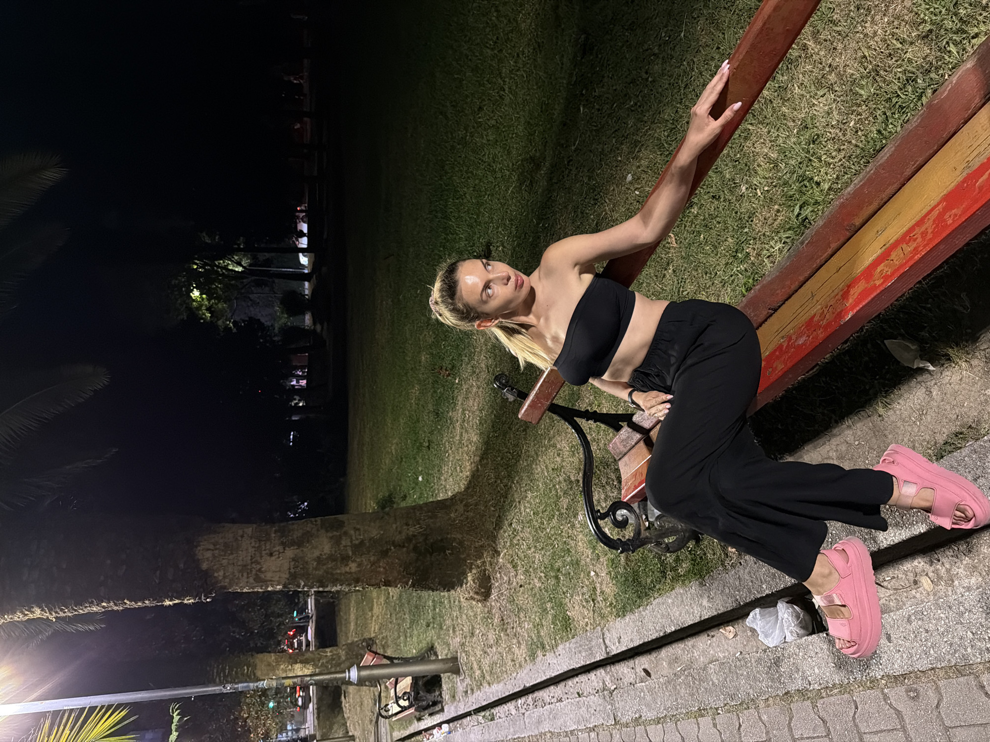 In Budva, Montenegro, at night, a blonde woman with her hair in a ponytail sits on a park bench. She is wearing a black bandeau top and black wide-leg pants. Her pink platform sandals are visible. The bench has a black metal frame and a wooden seat with red and yellow painted slats. She is sitting to the right of the bench, with her left hand resting on the armrest and her right leg crossed over her left. The bench is situated on a grassy area next to a paved walkway. A palm tree trunk is visible to the left of the woman, and another bench can be seen further down the walkway. Streetlights illuminate the scene, casting shadows on the grass and pavement. In the background, trees and buildings are visible in the darkness.