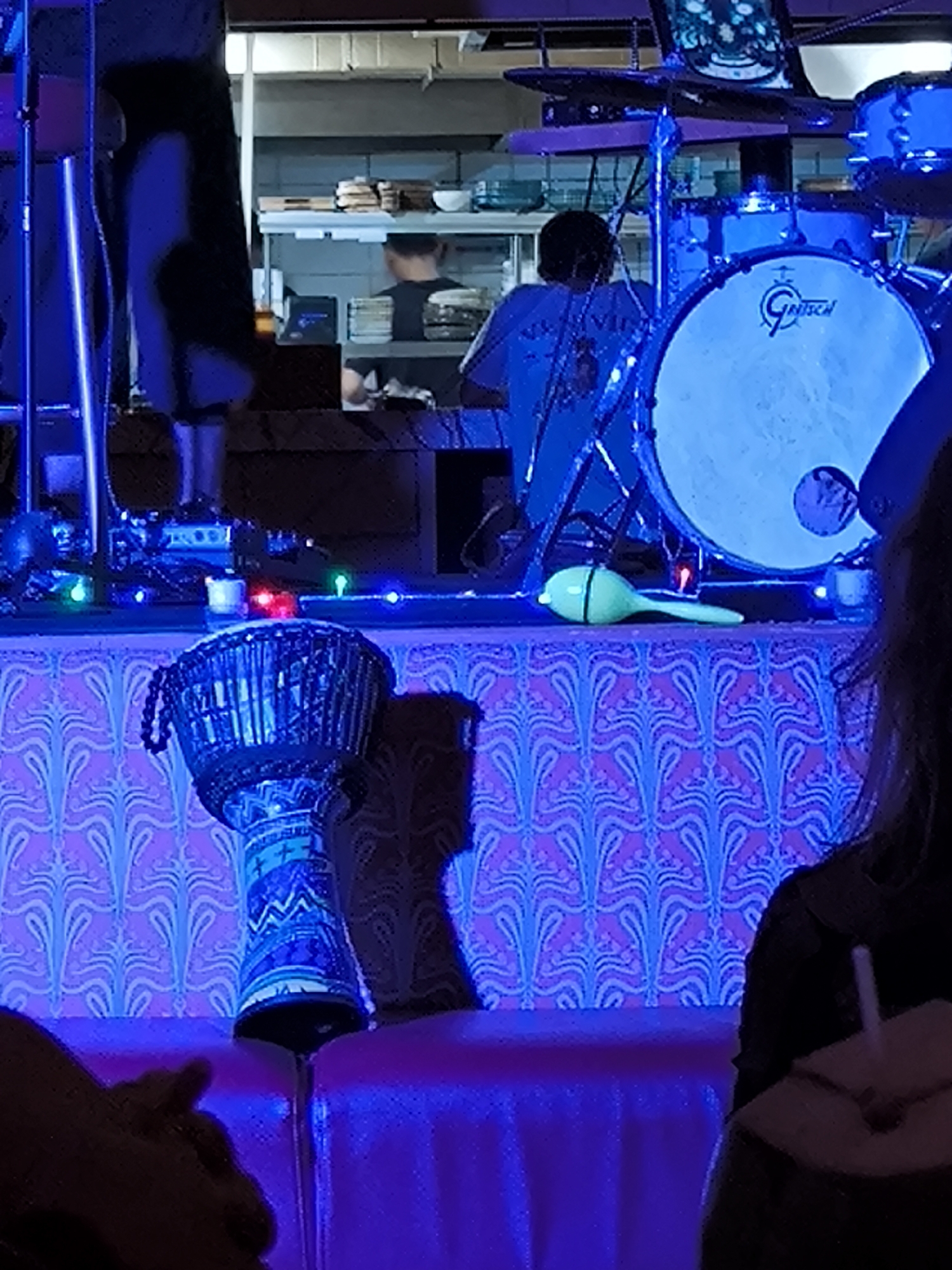 Captured under a blue lighting scheme, the image reveals a scene in North Kuta, Indonesia, potentially a live music venue or event space. A drummer is positioned behind a drum set. In the foreground is a djembe drum. A maraca is near the drum set. Behind the band, a kitchen area is visible with at least one person in the kitchen.
A woman with shoulder length hair is drinking from a coconut.
