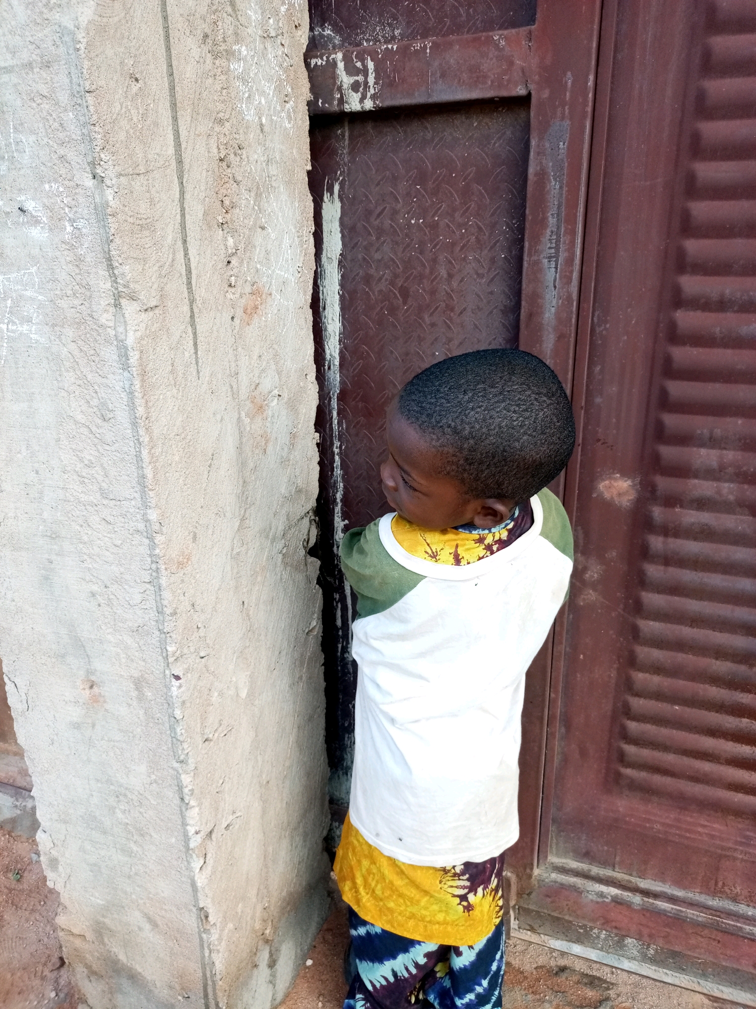 A young Black child, facing away from the camera and looking to their left, stands in front of a weathered, red-brown metal door with horizontal slats. The child wears a white t-shirt with green sleeves, a yellow undershirt, and patterned pants. To the left of the child is a rough, textured concrete pillar. The scene appears to be outdoors, possibly in a village or urban setting in Tempure, Nigeria, given the building materials and the child's attire. The ground is sandy and dirt.