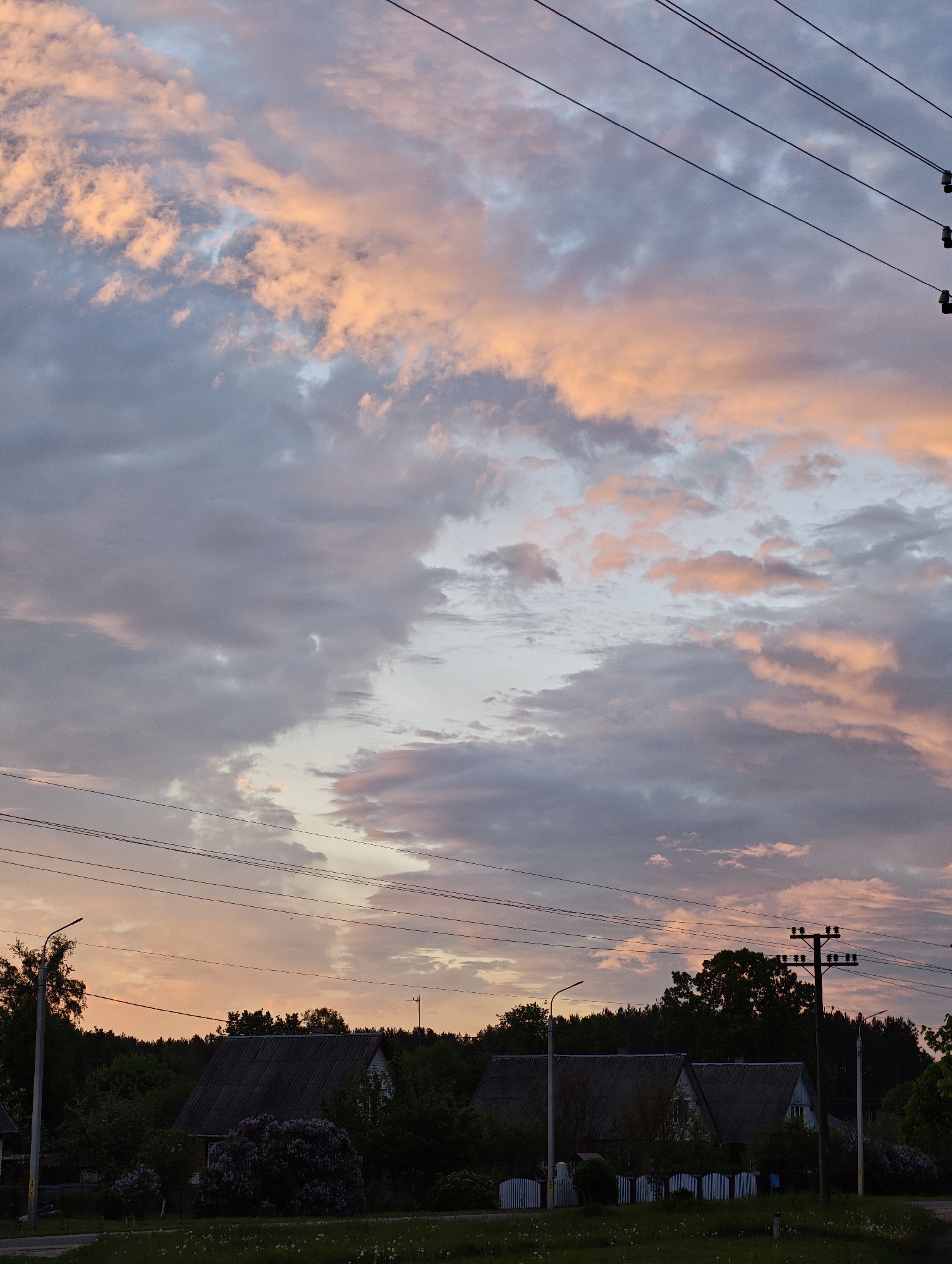 The image depicts a landscape at dusk or dawn, possibly in a rural or suburban area. The sky is filled with clouds, some tinged with pink and orange hues, suggesting the presence of a sunrise or sunset. Below the sky, the landscape is comprised of silhouettes of trees and houses with dark roofs. Power lines stretch across the top portion of the frame. Streetlights are visible amongst the houses, as well as what appears to be a utility pole. A white fence is partially visible in front of the houses. Lilacs are also visible. The specific location is unknown.