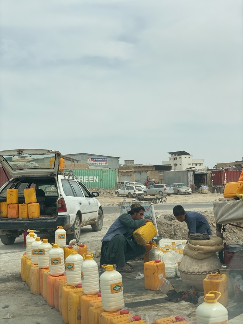 This outdoor scene captures an informal market or roadside stall in Kandahar, Afghanistan, under an overcast sky during daylight hours. Two individuals are engaged in the process of transferring and selling liquids. In the foreground, a man wearing traditional clothing and a hat squats, carefully pouring a liquid from a large yellow plastic jerry can into a smaller container on the ground. To his right, a younger individual leans over, seemingly attending to other containers.
A white hatchback car is parked to the left with its trunk open, revealing a stack of yellow jerry cans inside, some bearing unreadable Farsi/Dari script. In front of the car and extending across the foreground, numerous identical yellow jerry cans are neatly lined up on the ground, serving as a display stand. On top of these, and also on the ground, are various white plastic bottles and jugs of different sizes, filled with a white liquid. Many of these white containers feature a distinct green and white label, with "Crown" or similar text visible on some. A large, possibly clay or fabric, pot sits on the ground near the squatting man.
The background shows a dusty, semi-urban environment with multiple parked vehicles, including other cars and a bicycle, and a series of buildings. Large green shipping containers are prominent in the middle ground, one clearly marked with the word "GREEN." The scene portrays a practical, manual trading operation for essential commodities in a bustling setting.