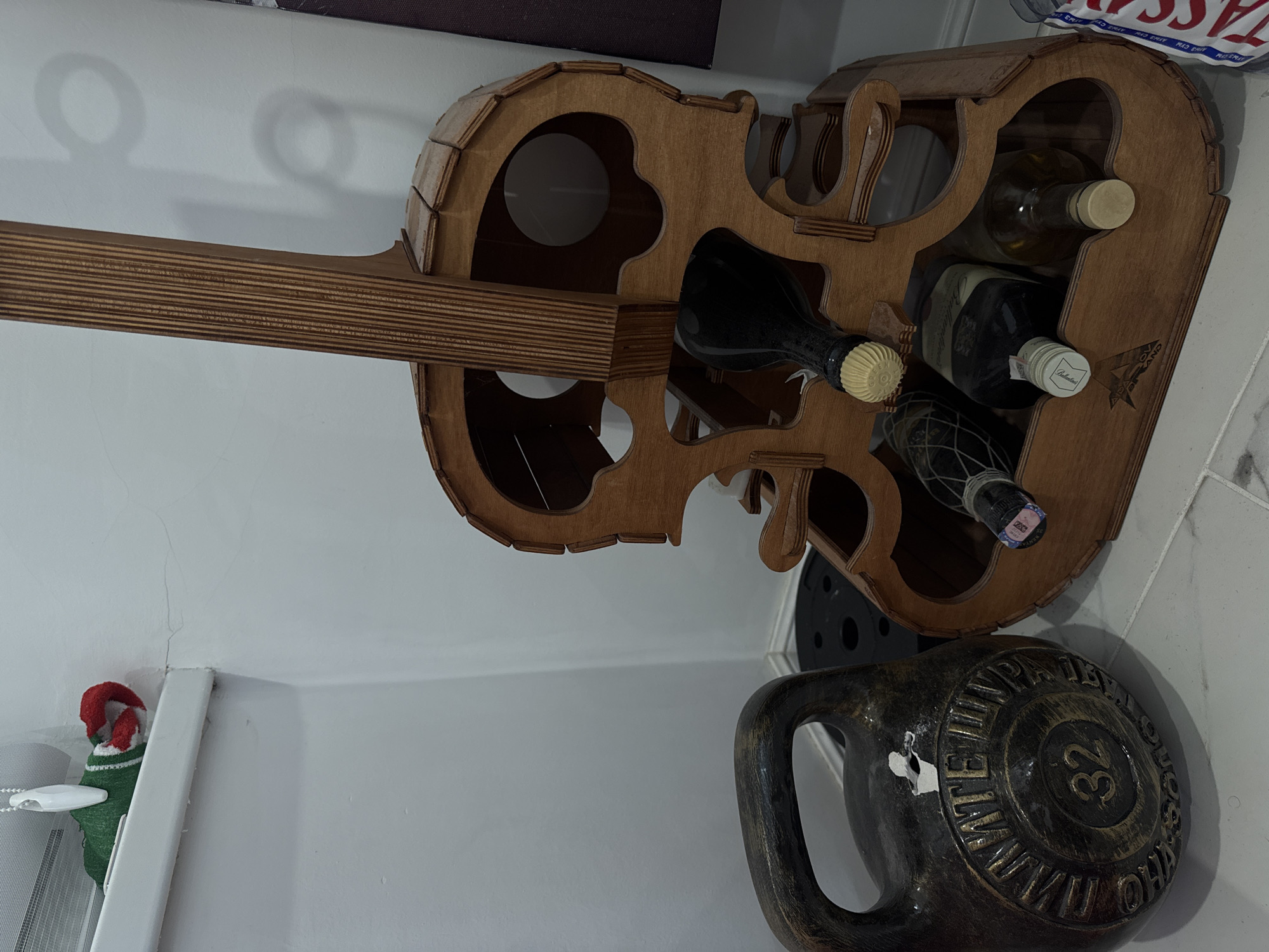 The image shows a dark brown kettlebell with the number "32" visible on its side, placed on a light-colored tiled floor. To the right of the kettlebell is a wooden wine rack shaped like a guitar, holding several bottles of wine. In the background, a white wall is visible, with a white blind partially drawn at the top left, where a green and red striped sock is draped over the edge. The scene appears to be indoors, possibly in a room or storage area. There is no indication of the city of Oral, Kazakhstan.