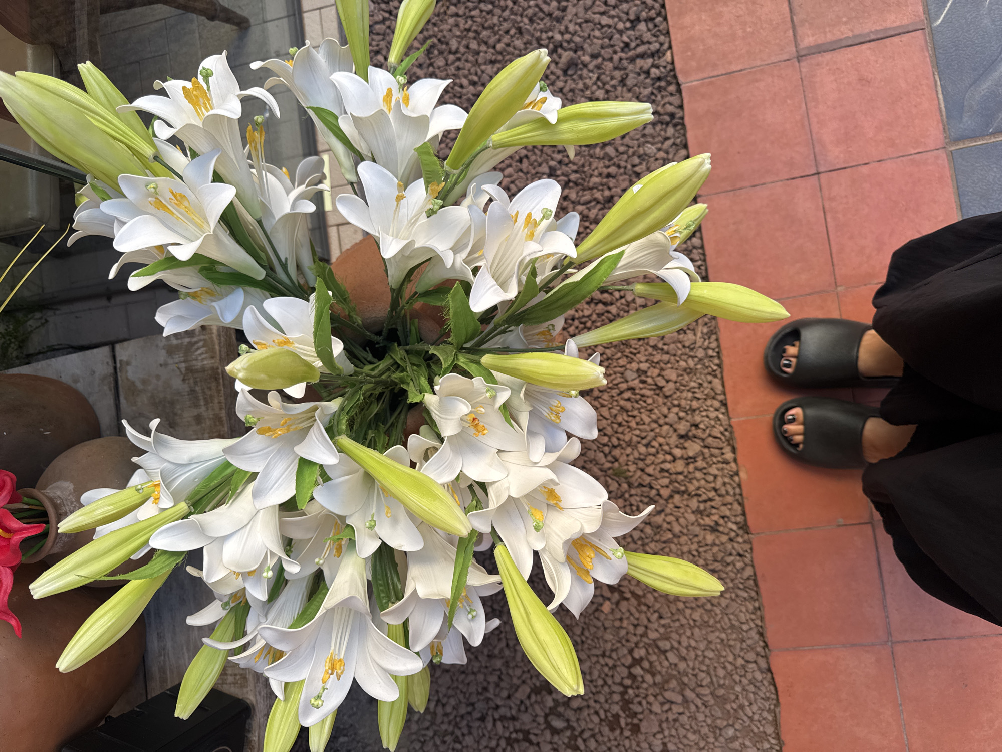 The scene, located in Nha Trang, Vietnam, presents an overhead view centered on a large bouquet of white lilies. The bouquet features numerous fully open white blossoms, each displaying prominent yellow anthers and stigmas, intermingled with several green, unopened lily buds. The lower legs and feet of one individual are visible in the foreground, wearing black wide-leg trousers and dark slip-on sandals; their toenails are painted dark. The person stands on a reddish-brown tiled surface that transitions to a dark brown gravel-textured area on the right. In the upper background, blurred, multiple terracotta pots are visible, one containing red artificial floral elements. The environment appears to be an outdoor or semi-outdoor setting under natural illumination.