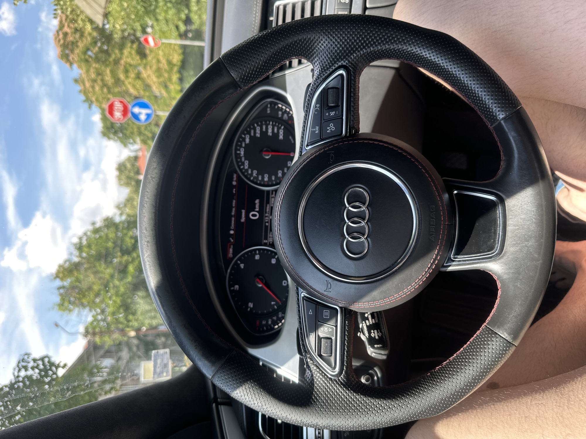 The image shows a close-up view of the steering wheel of an Audi vehicle, likely taken from the driver's seat. The steering wheel is black leather with red stitching and the Audi logo is prominently displayed in the center. The dashboard instruments, including a speedometer showing 0 km/h, are visible behind the steering wheel. Traffic signs, including a stop sign and a directional sign, are visible through the windshield, suggesting the car is in a street environment. Trees and buildings are also visible in the background, indicating an urban setting. Based on the specific design of the traffic signs (a red octagonal stop sign with white lettering, and a blue circular sign with a white arrow indicating straight ahead and left turn), and the general architectural style of the buildings visible, this scene is consistent with a location in Ruse, Bulgaria. The image captures a moment of stillness, possibly at a traffic light or stop sign.