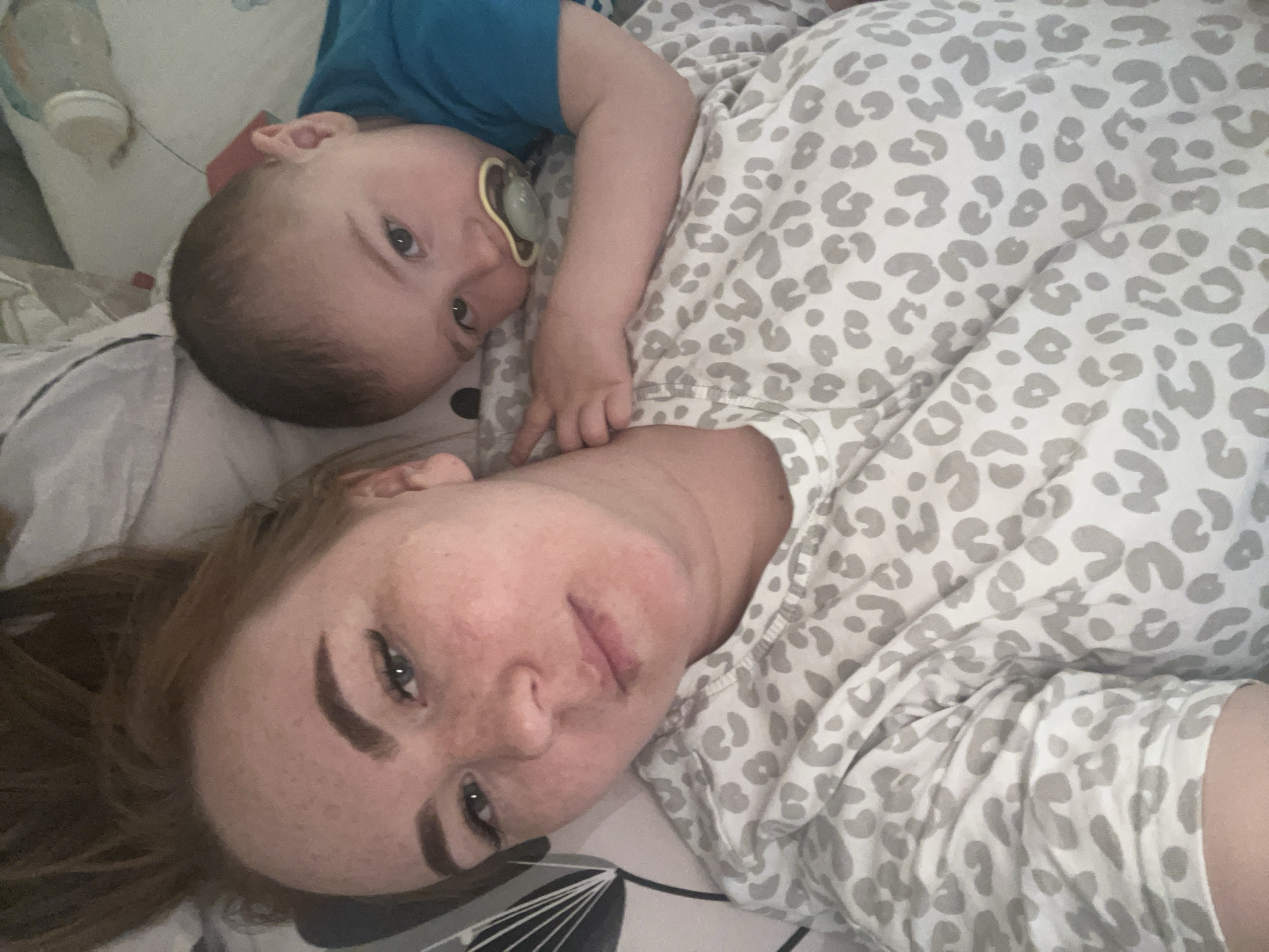 A woman and a baby are lying on a bed. The woman has red hair and is wearing a white shirt with gray leopard spots. The baby is wearing a blue shirt and has a pacifier in his mouth. A baby bottle is on the bed beside the baby. The location is likely indoors in a residential setting. There is no evidence to support the claim that the image was taken in Chaikovsky, Russia.