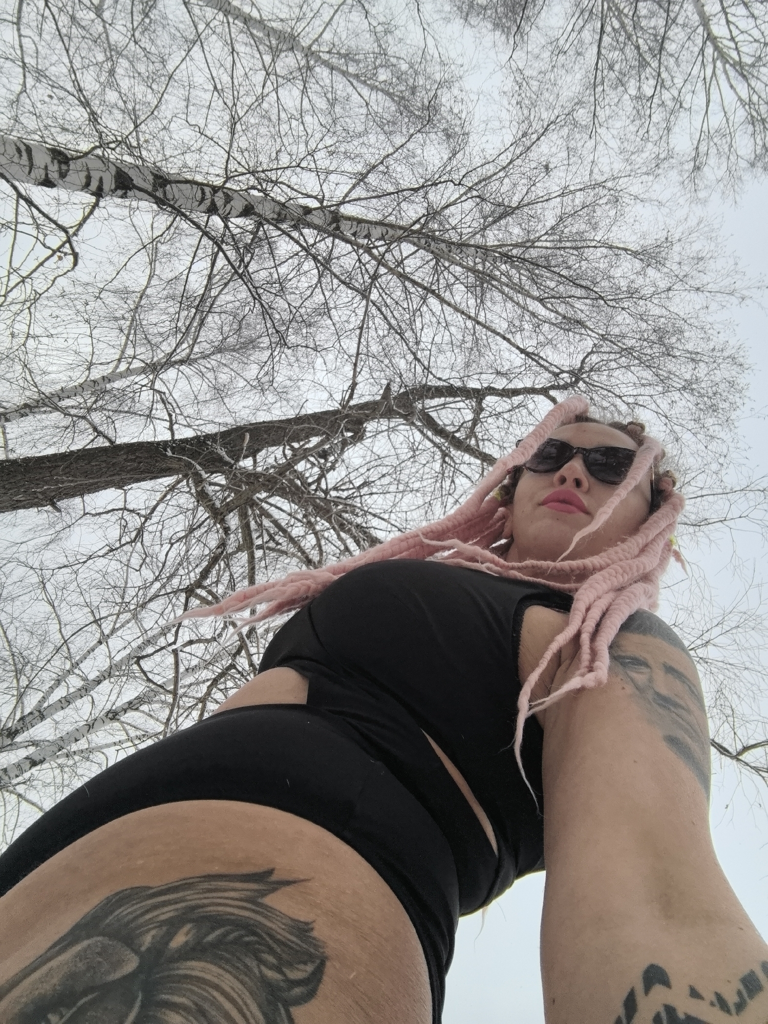 A woman with pink dreadlocks and sunglasses is looking upwards towards the camera. She is wearing a black top and leggings. She has tattoos on her left thigh and right arm. The background consists of bare birch trees against a grey sky, suggesting a winter scene. The location is likely in a wooded area of Noviy, Russia.