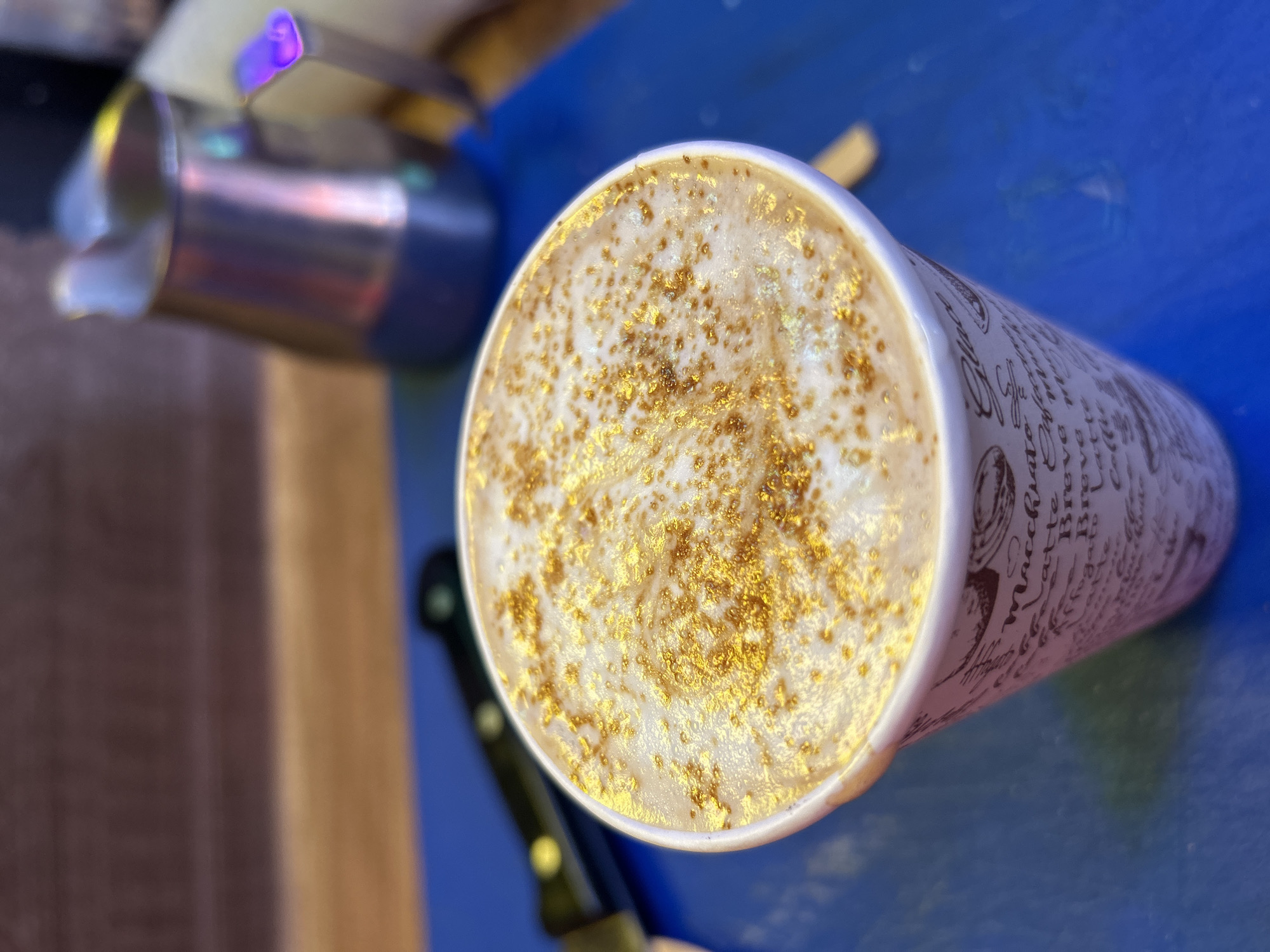 A close-up, high-angle shot shows a paper coffee cup filled with a frothy beverage. The beverage is topped with a light brown foam and sprinkled with gold glitter. The cup has a patterned design featuring various coffee-related words in brown lettering. In the background, out of focus, is a stainless steel milk pitcher with a purple handle, and a black-handled knife. The objects are resting on a bright blue surface, with a wooden surface in the blurred background. The scene appears to be in a cafe or coffee shop.