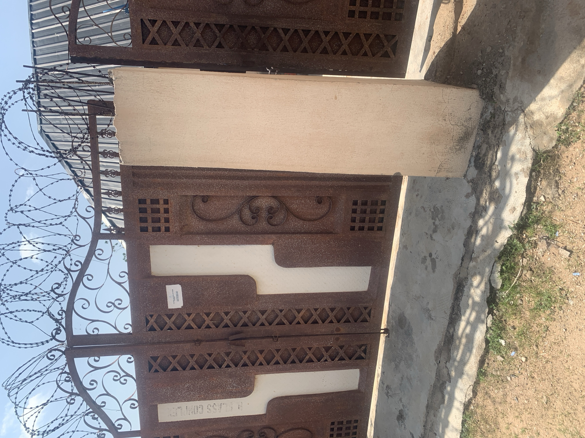 The image shows a close-up of a rusty metal gate with ornate scrollwork and diamond-shaped cutouts. Barbed wire is wrapped around the top of the gate, suggesting a security-conscious environment. The gate appears to be part of a larger structure, possibly a building or compound.
The gate is divided into two sections, with a horizontal latch mechanism visible in the middle. The metal has a weathered, oxidized appearance, with patches of rust and some lighter-colored paint or material applied in horizontal bands across the gate's surface. The bottom section of the right-hand gate has a label with white text on a dark background. The text, which is partially obscured and appears to be reversed, reads "CLASS COMPLEX".
The background is a mix of light-colored concrete and dry, sandy ground with sparse green vegetation. The sky is visible through the openings in the gate, and it appears to be a clear day. The lighting suggests it is daytime, with shadows cast across the concrete. The overall impression is one of a utilitarian structure that has seen some wear and tear, located in a warm, possibly arid environment. There are no people visible in the frame.