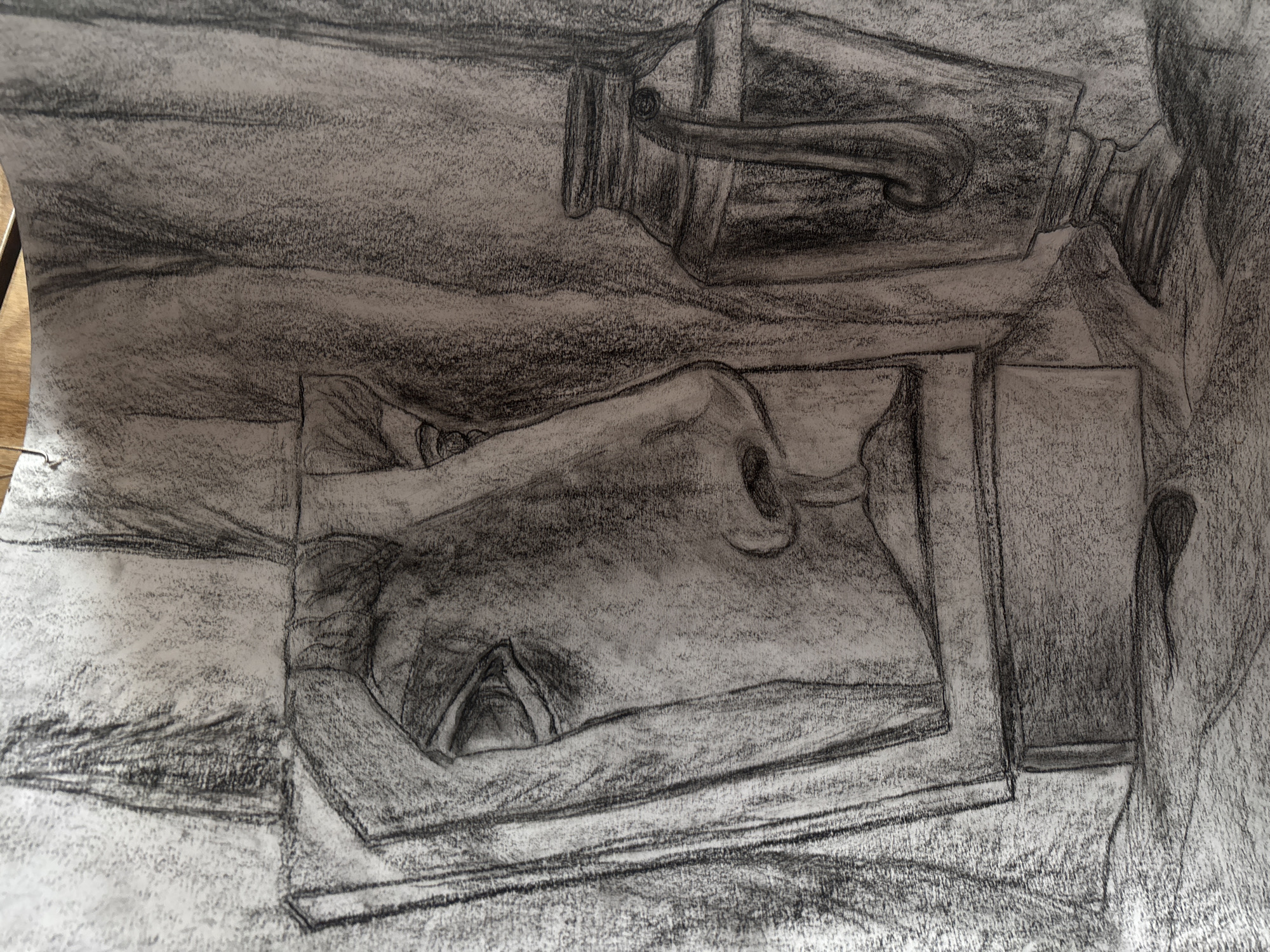 A charcoal drawing depicts a still life composition. The primary subjects are two perfume bottles, one lying horizontally and the other vertically, placed on a surface with a rectangular cutout. The background consists of a textured, charcoal-shaded surface. The scene seems to be drawn from an elevated, slightly angled perspective. The drawing is not specifically identifiable to the city of Troyan, Bulgaria.