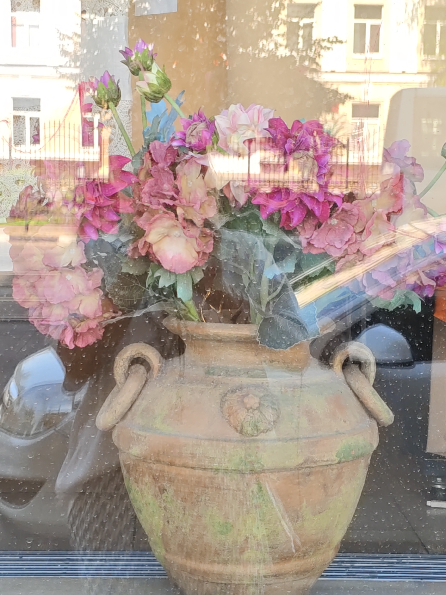 The image shows a close-up, slightly distorted view of a terracotta amphora filled with a bouquet of artificial flowers. The amphora is decorated with raised circular handles and a relief of a rosette. It exhibits signs of age and weathering, with patches of green moss growing on its surface. The flowers are a mix of pink and purple hues, with some buds still closed and others in full bloom. The bouquet is lush and full, with green artificial foliage. The entire scene is viewed through a glass surface that has water droplets on it, creating a reflective and somewhat obscured view. In the background, through the glass, reflections of buildings and trees are visible, suggesting an urban environment. The light suggests it is daytime, possibly with sunlight casting shadows. This scene is located in Rostov-on-Don, Russia.