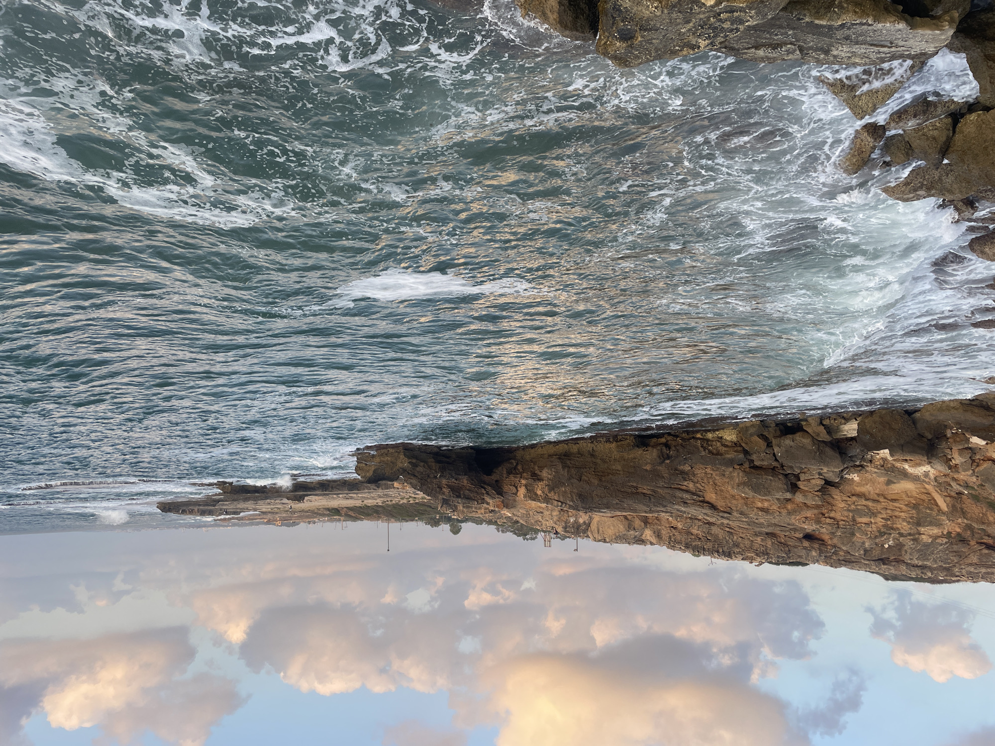 The image depicts a rocky coastline in the Mate Asher Regional Council, Israel, likely near Achziv National Park. The sea is a deep turquoise with whitecaps and foam from the waves crashing against the rocks and the shore. The sky is a pale blue with scattered clouds tinged with the warm hues of sunset. Jagged, brown rocks dominate the left side of the image, forming a rugged cliff face and leading down to the water. In the distance, the coastline continues with more rock formations and a hint of a town or structures further along the shore. The overall scene is one of natural coastal beauty under a dynamic sky.